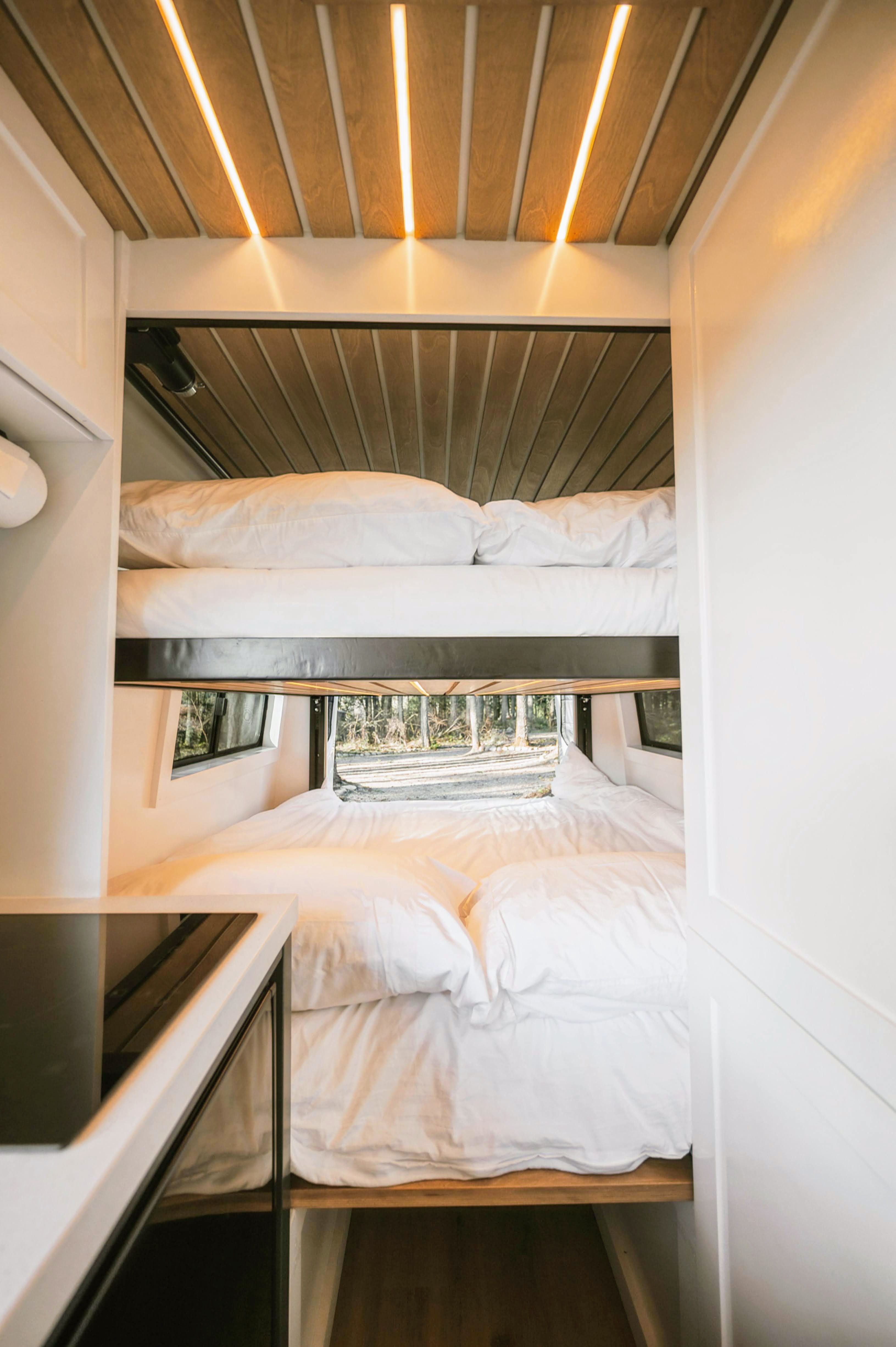 Inside a modern RV rental: stacked bunk beds with white linens, ambient wood ceiling lighting, and a window view of a serene forest.. Mercedes-Benz Sprinter 2021