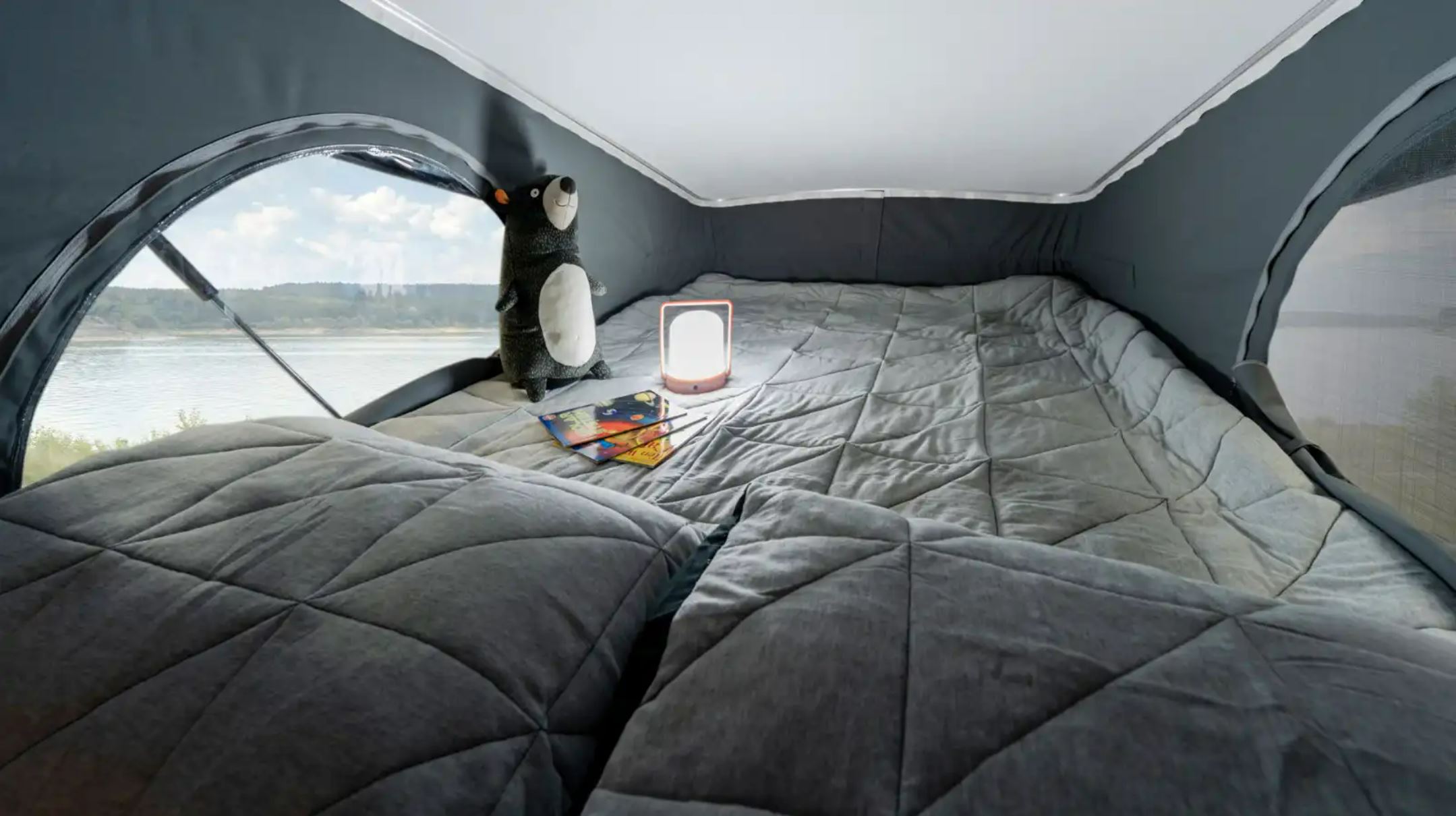 RV rental's cozy pop-up bed offers a tranquil lake view. Complete with grey quilted bedding, lamp, bear, and books.. Winnebago Solis 2023