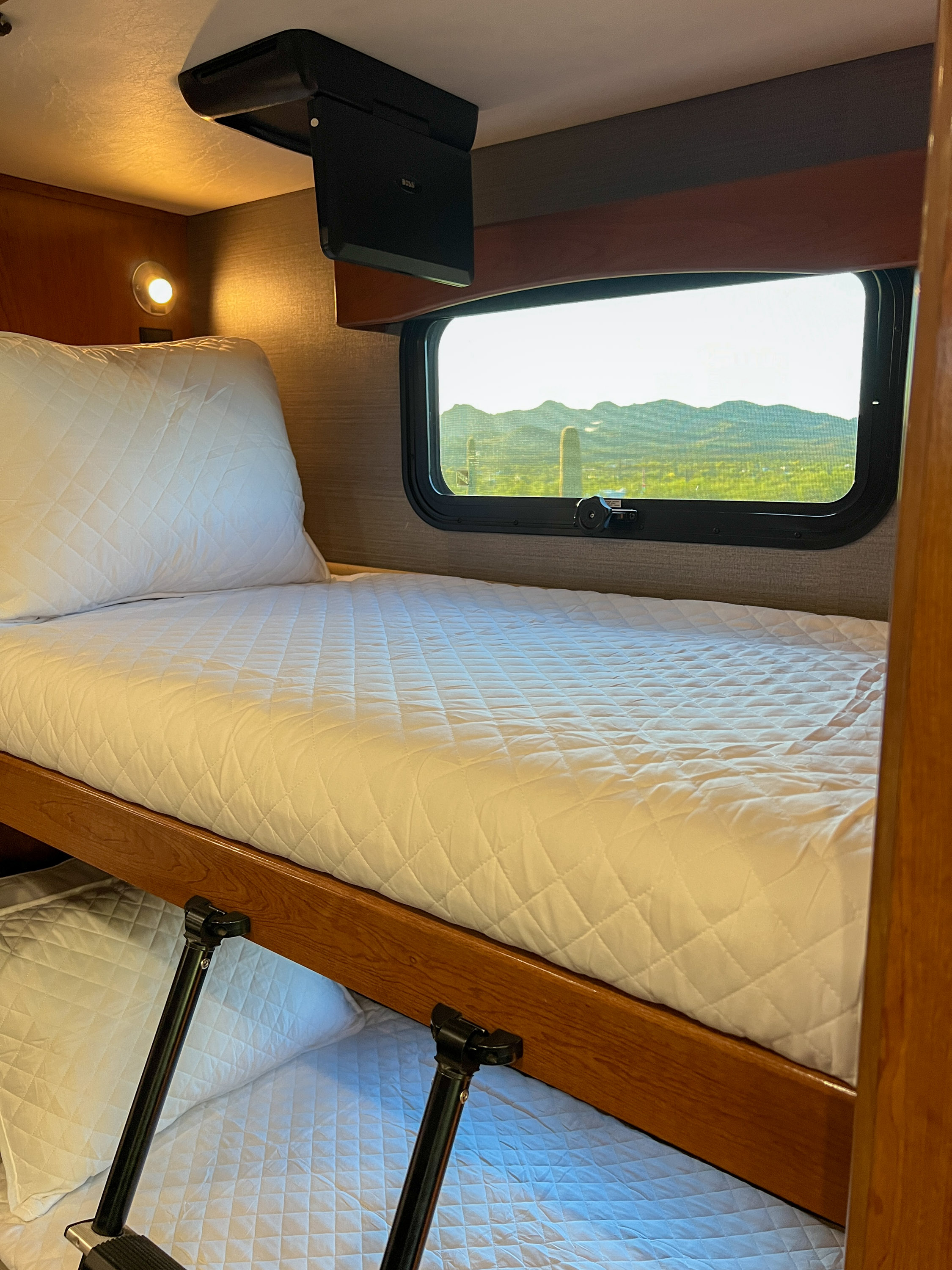 Cozy RV rental bunk bed area, featuring quilted bedding, a ceiling TV, and a window with a stunning desert mountain view.. Fleetwood Discovery 2018