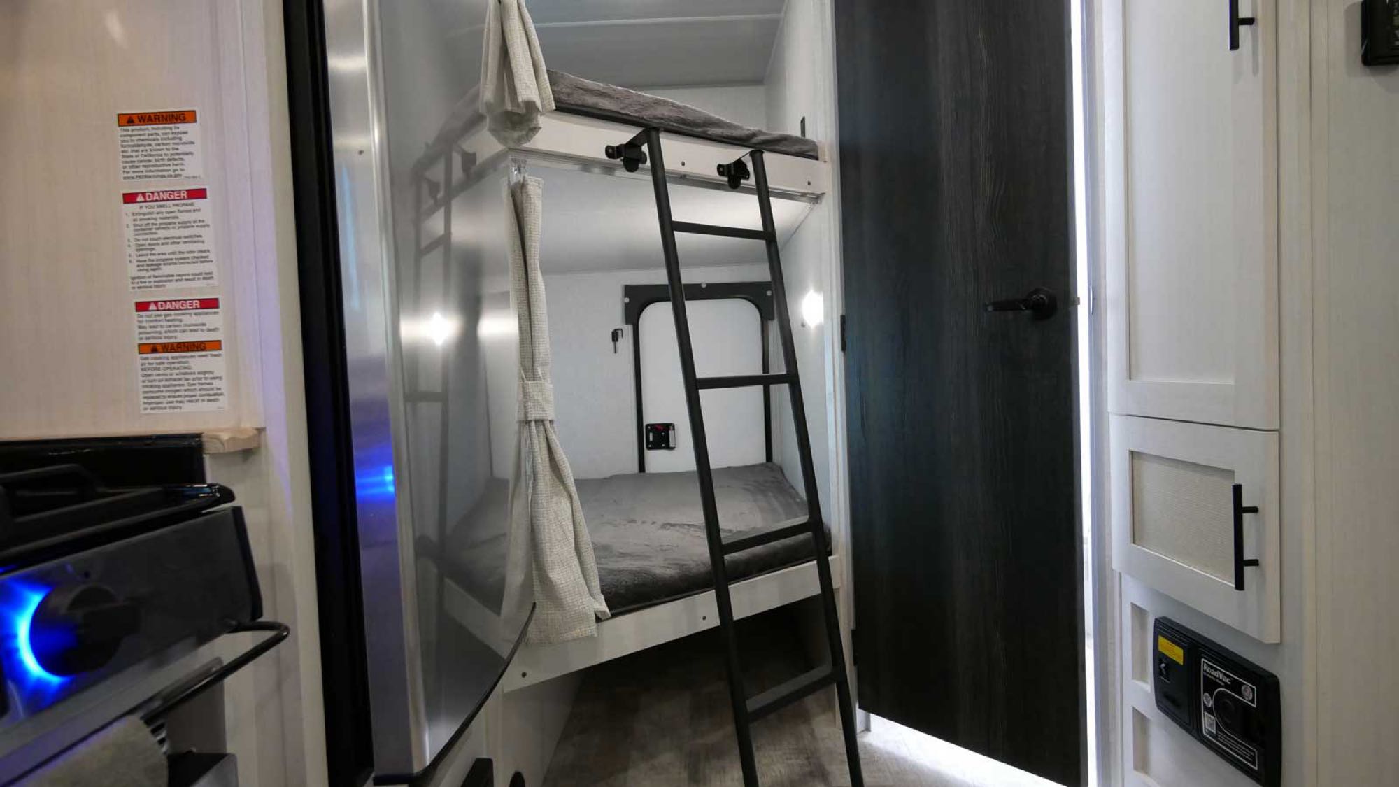 An RV rental interior showing bunk beds with ladder and privacy curtains, next to a compact kitchen area.. Forest River NB 20.3 2023