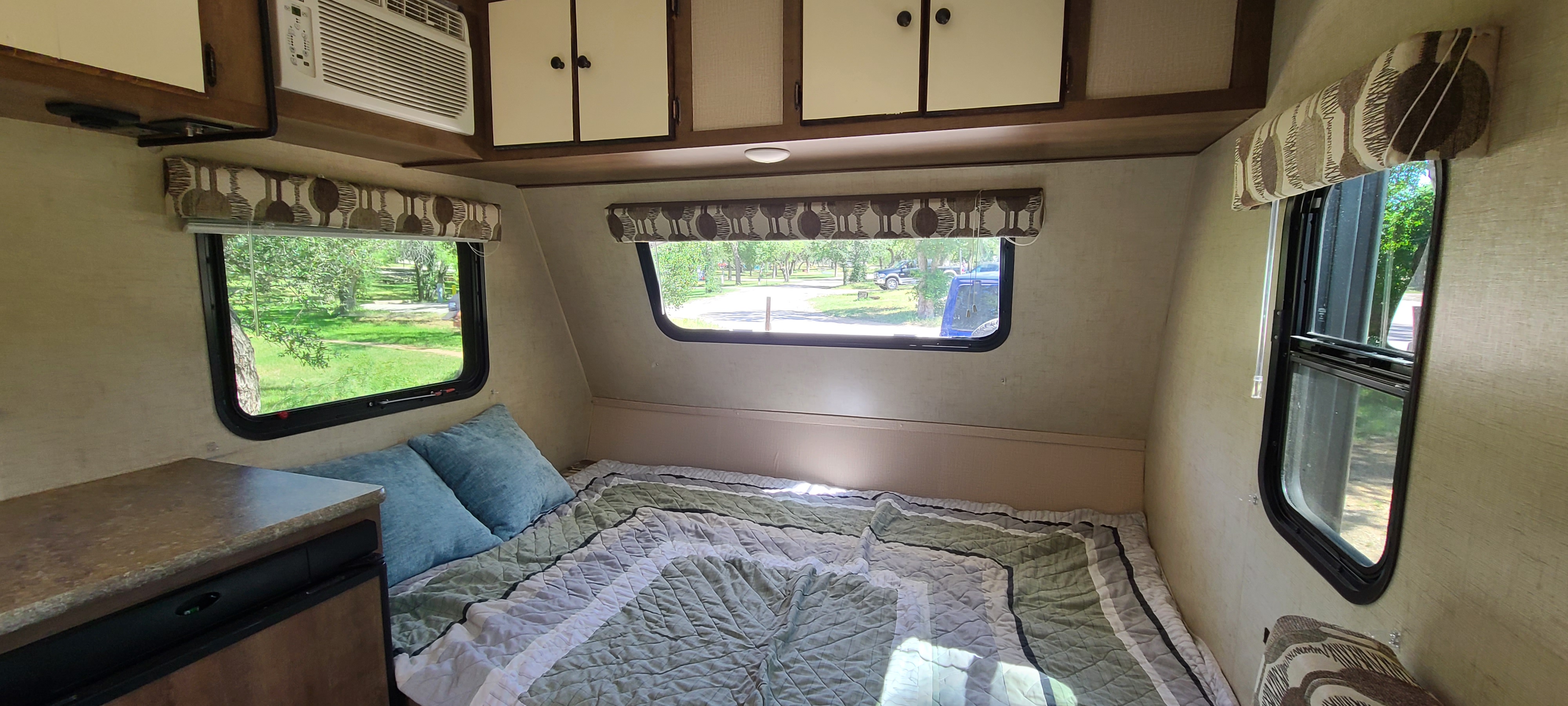 Inside a comfortable RV rental: A compact bedroom with a green quilt, blue pillows, and windows showing outdoor scenery.. KZ Sportsmen 2018