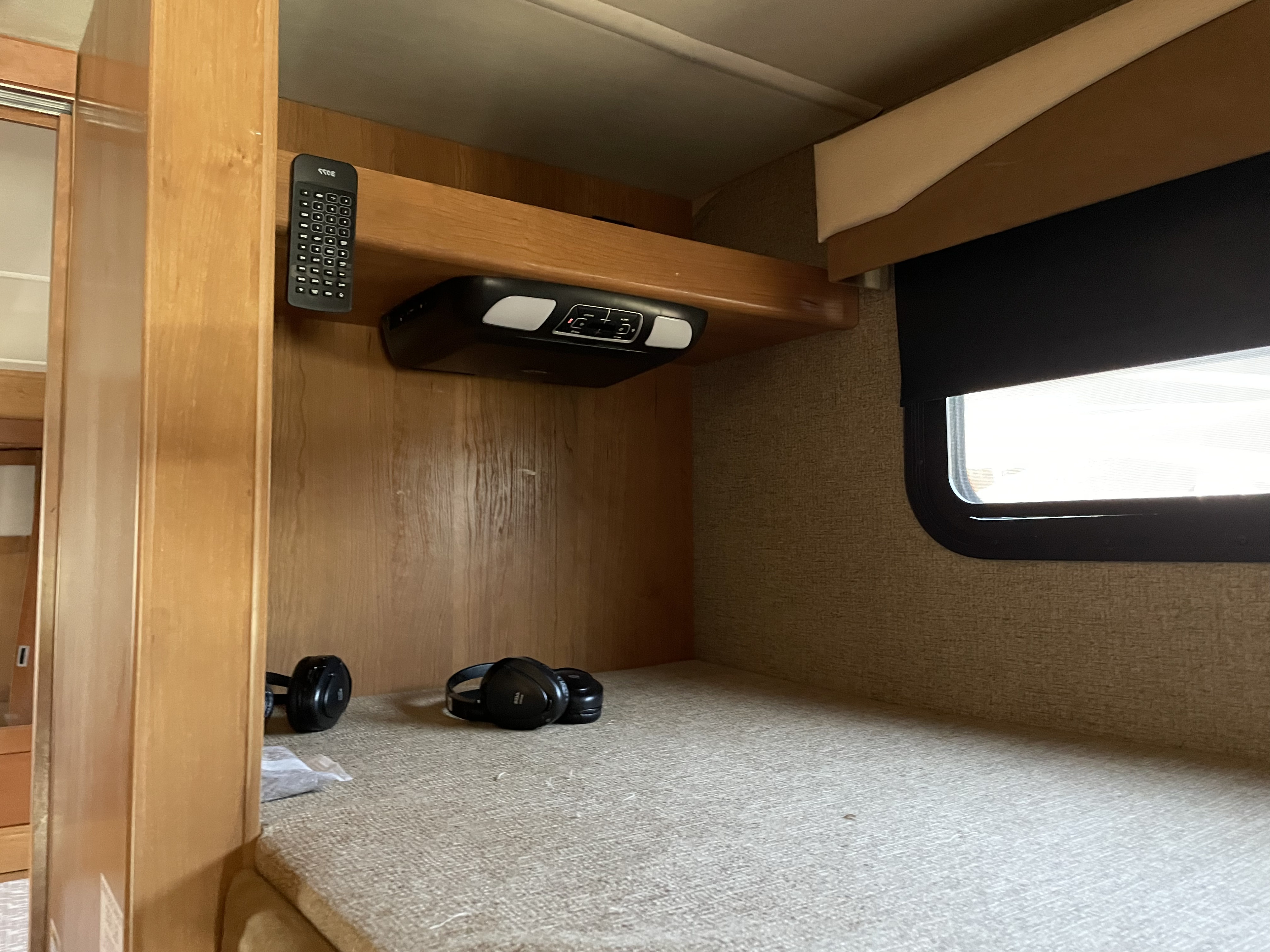 Interior of an RV rental showing a lower bunk bed, overhead entertainment system, window with shade, and headphones.. Fleetwood Excursion 2015