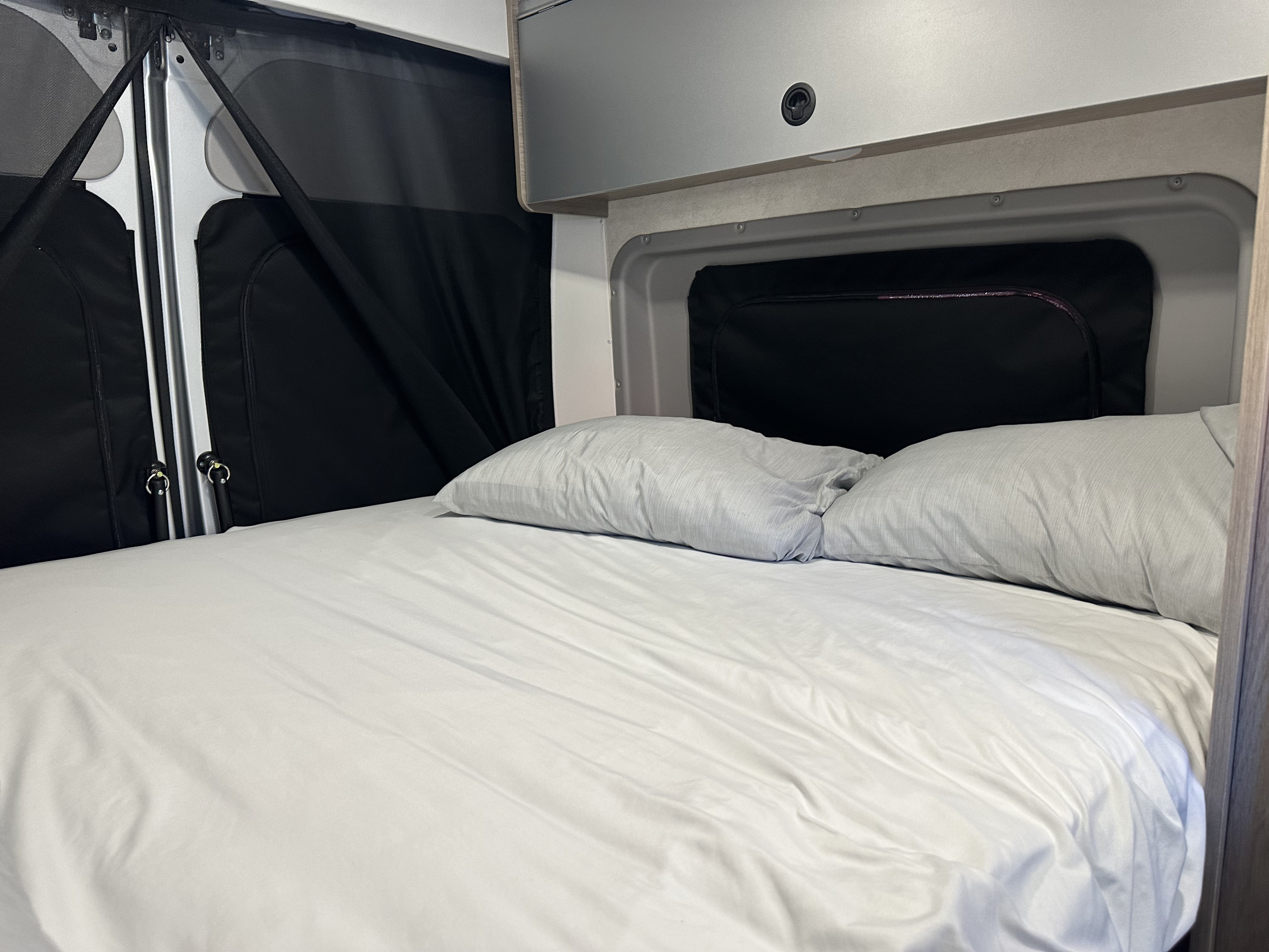 Comfortable **rv rental** bed with white sheets & grey pillows, featuring window screens and overhead cabinets.. 2023 Winnebage Ram Pro master 3500 2023