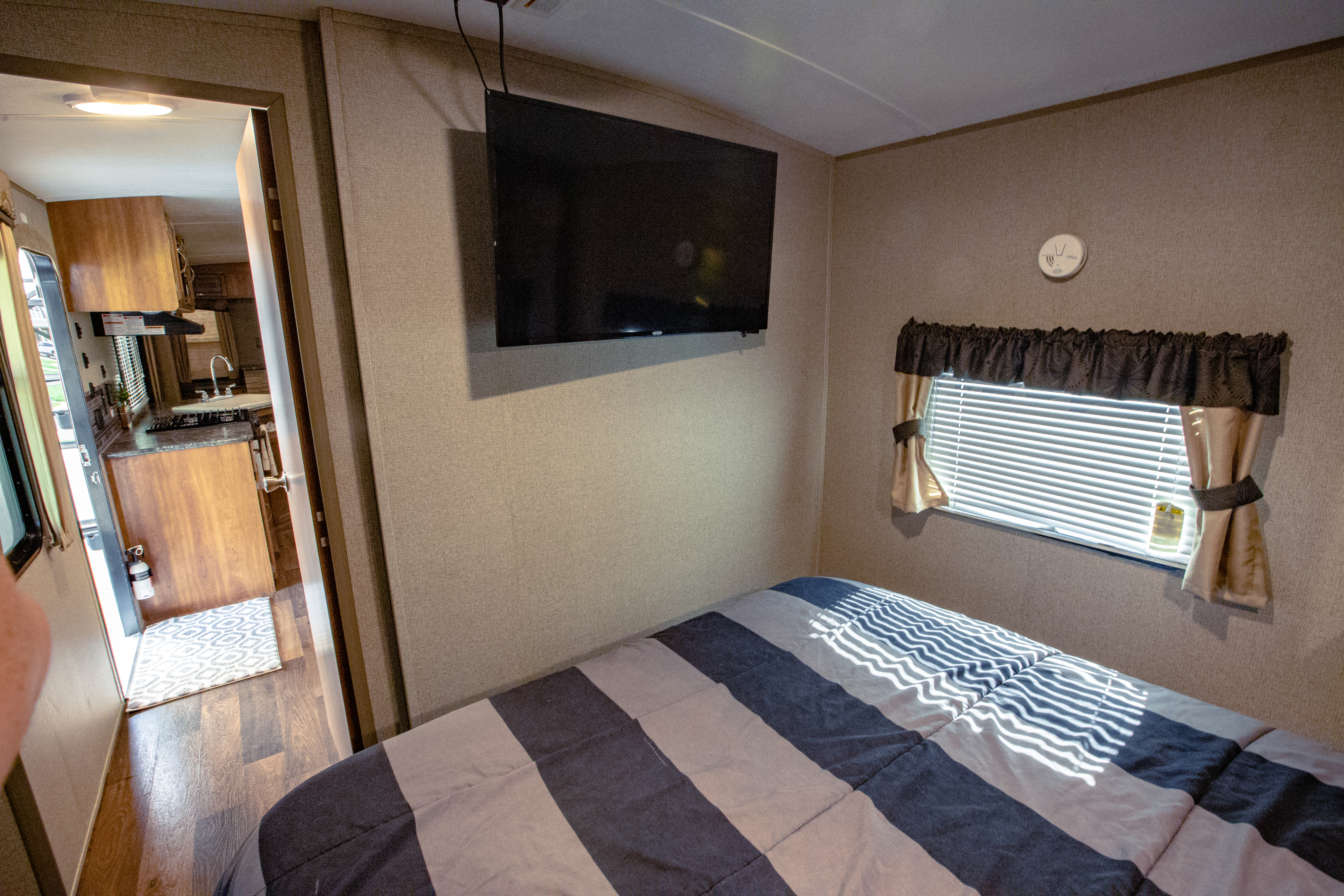 Cozy RV rental bedroom with a striped bed, mounted TV, window, and a peek into the functional kitchen area.. Keystone RV Springdale 2017