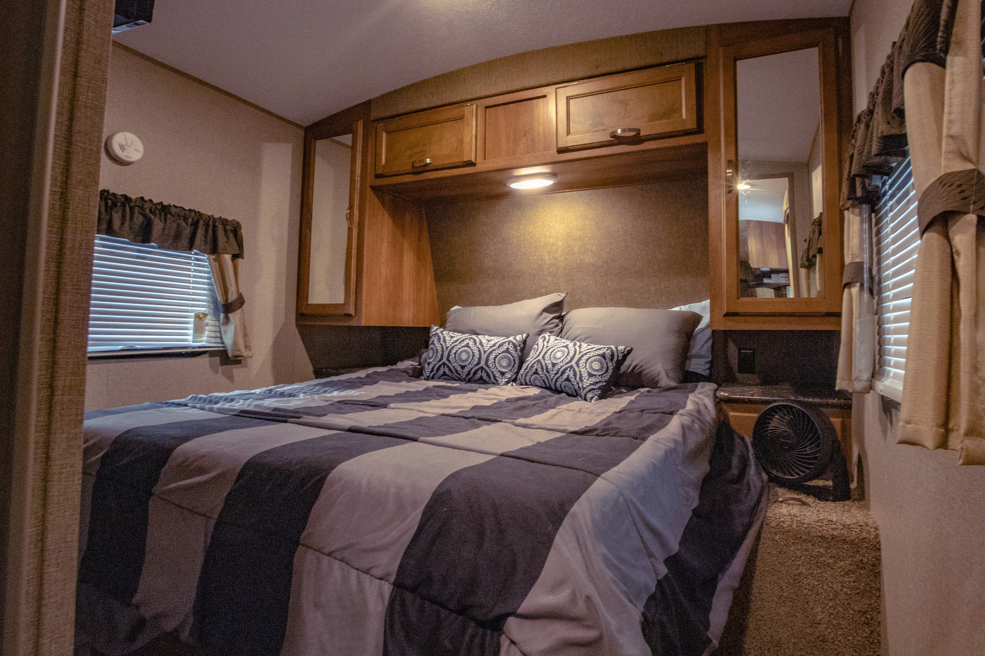Cozy RV rental bedroom features a striped bed, wooden cabinets, a window with blinds, and a small fan.. Keystone RV Springdale 2017