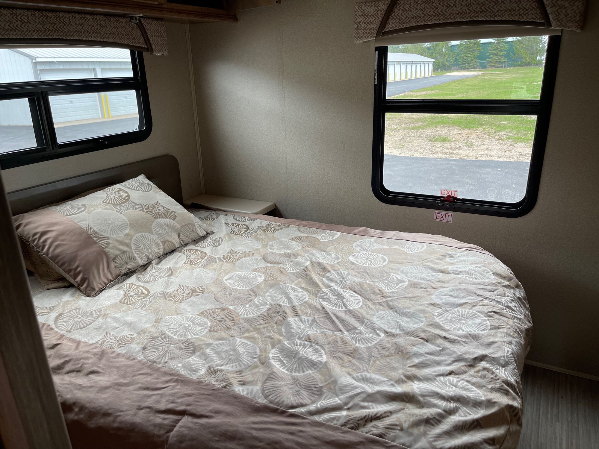 Cozy RV rental bedroom featuring a patterned comforter, small nightstand, and two windows with an outdoor view.. Jayco Greyhawk Prestige 2019