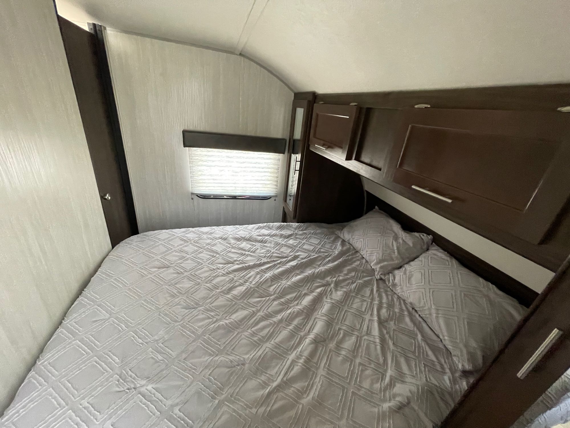 Comfortable RV rental bedroom featuring a grey bed, overhead storage, and a window with blinds.. Forest River Cherokee 2019