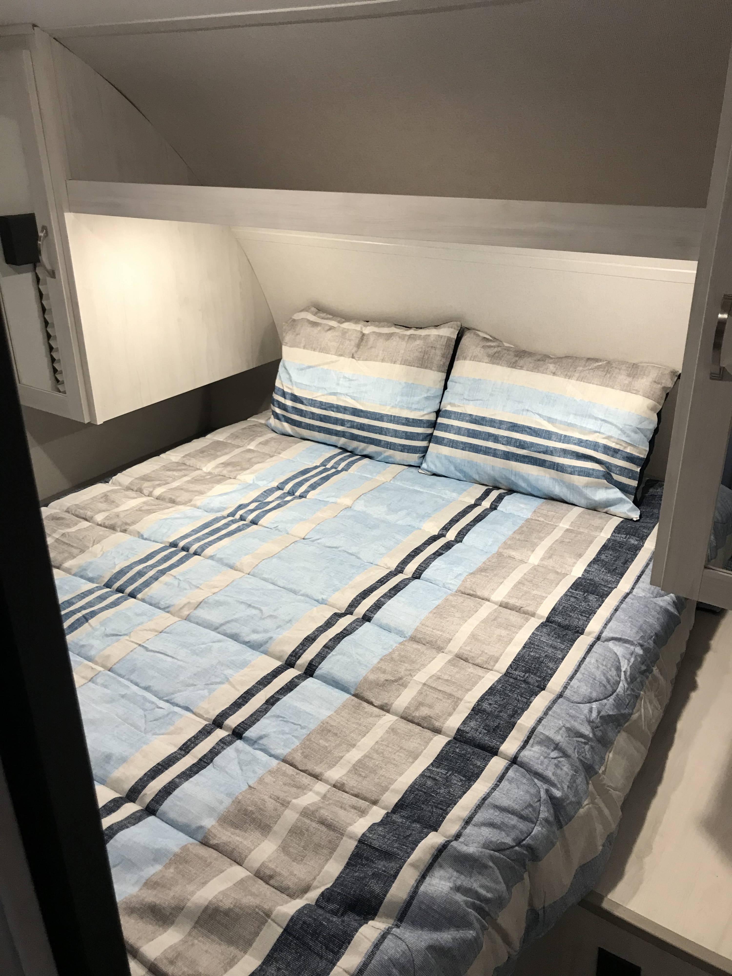 An RV rental bedroom featuring a bed with blue and beige striped bedding, pillows, and built-in overhead storage.. Forest River Tracer LE 2022