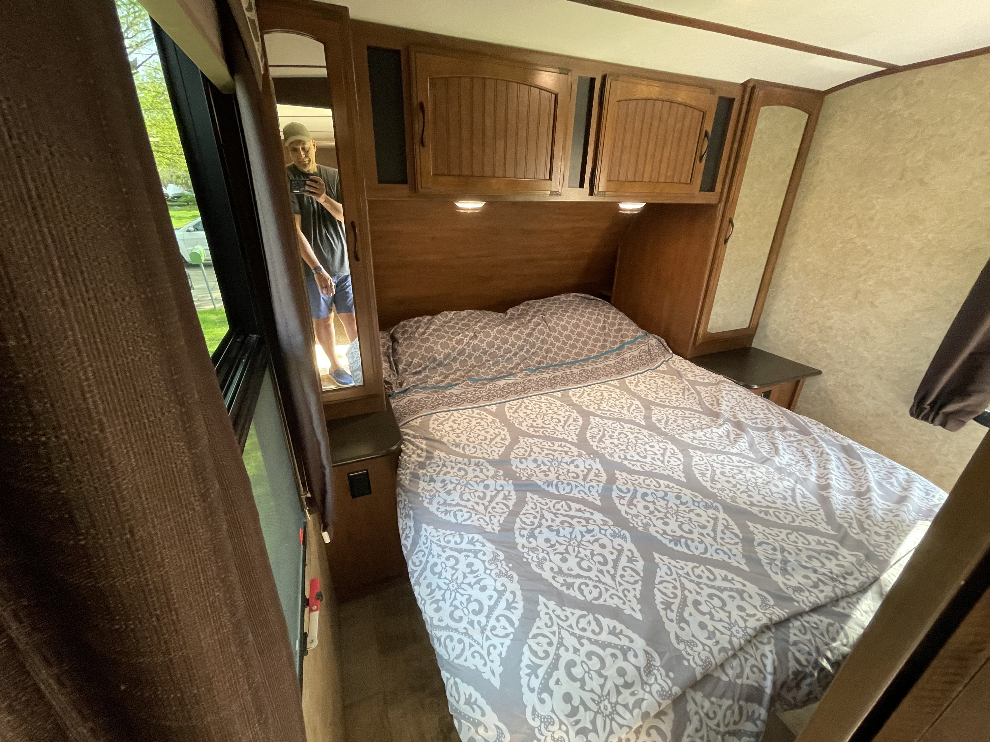 An RV rental bedroom: cozy bed with patterned bedding, overhead cabinets, and a mirror showing the photographer. Window view.. Jayco White Hawk 2017