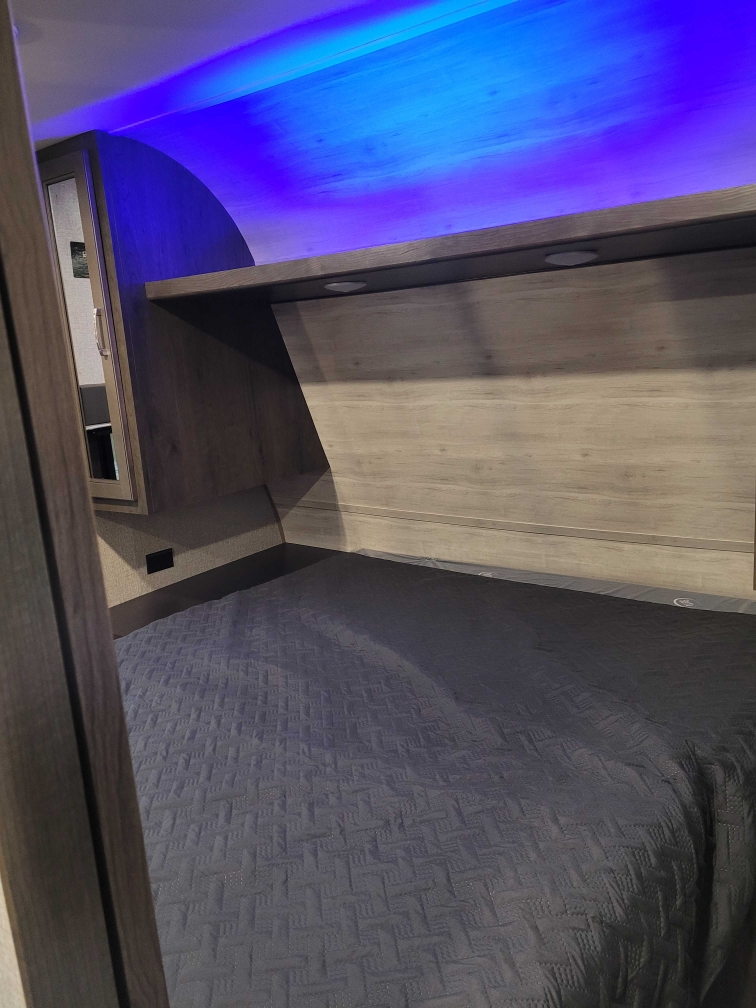 Modern RV rental bedroom with a dark quilted bedspread, overhead shelf, recessed lights, and vibrant blue LED ceiling glow.. Jayco Jay Flight SLX Rocky Mountain Edition 2021