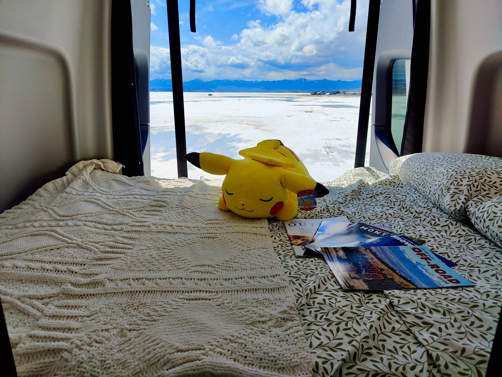 An RV rental offers a cozy bed view of the Bonneville Salt Flats, mountains, and sky. A Pikachu plush rests on the bed.. Winnebago Revel 2023