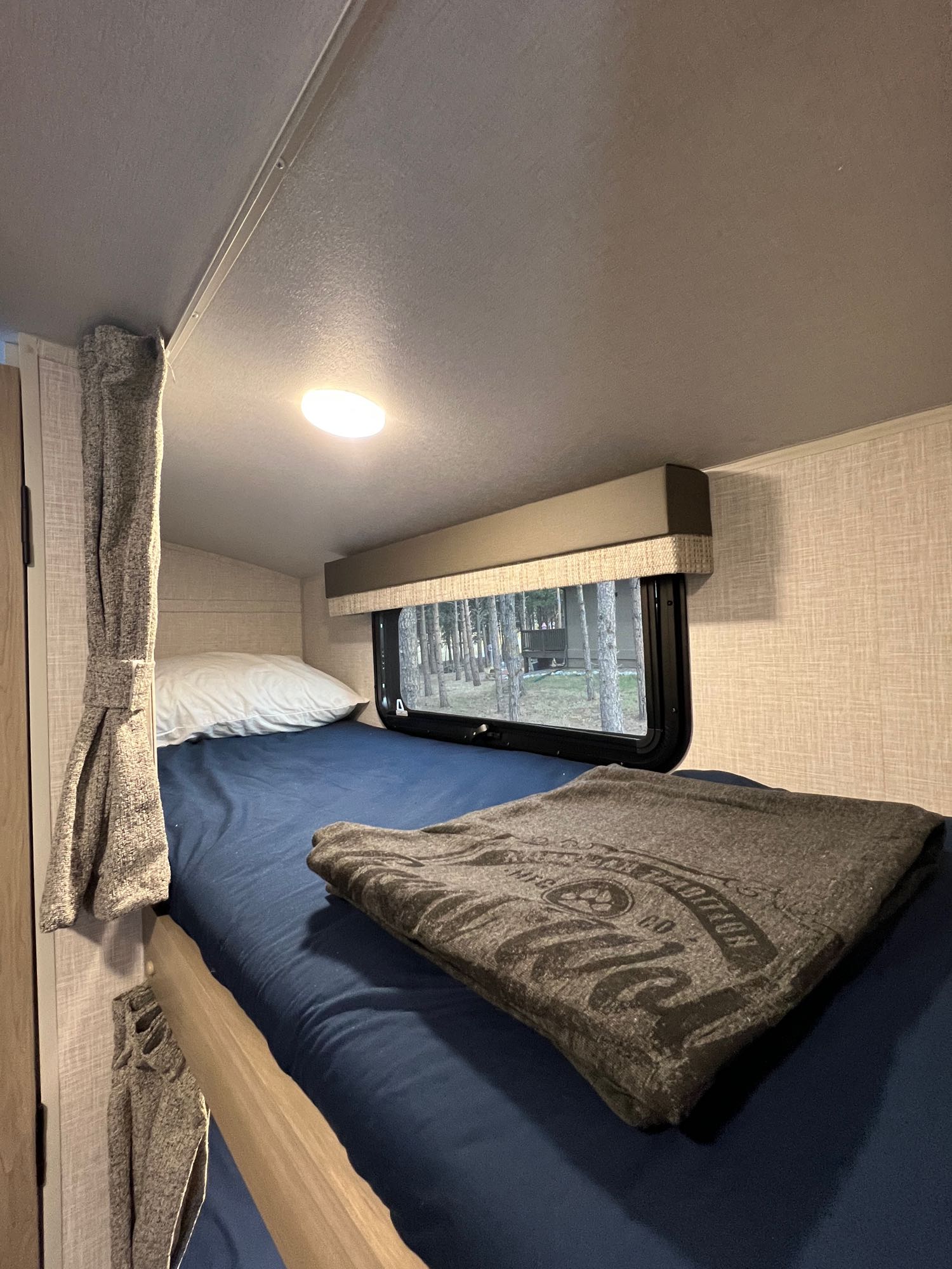 Cozy RV rental bunk bed interior with a blue mattress, white pillow, and folded blanket, offering a wooded view.. Forest River Flagstaff E-Pro 2023