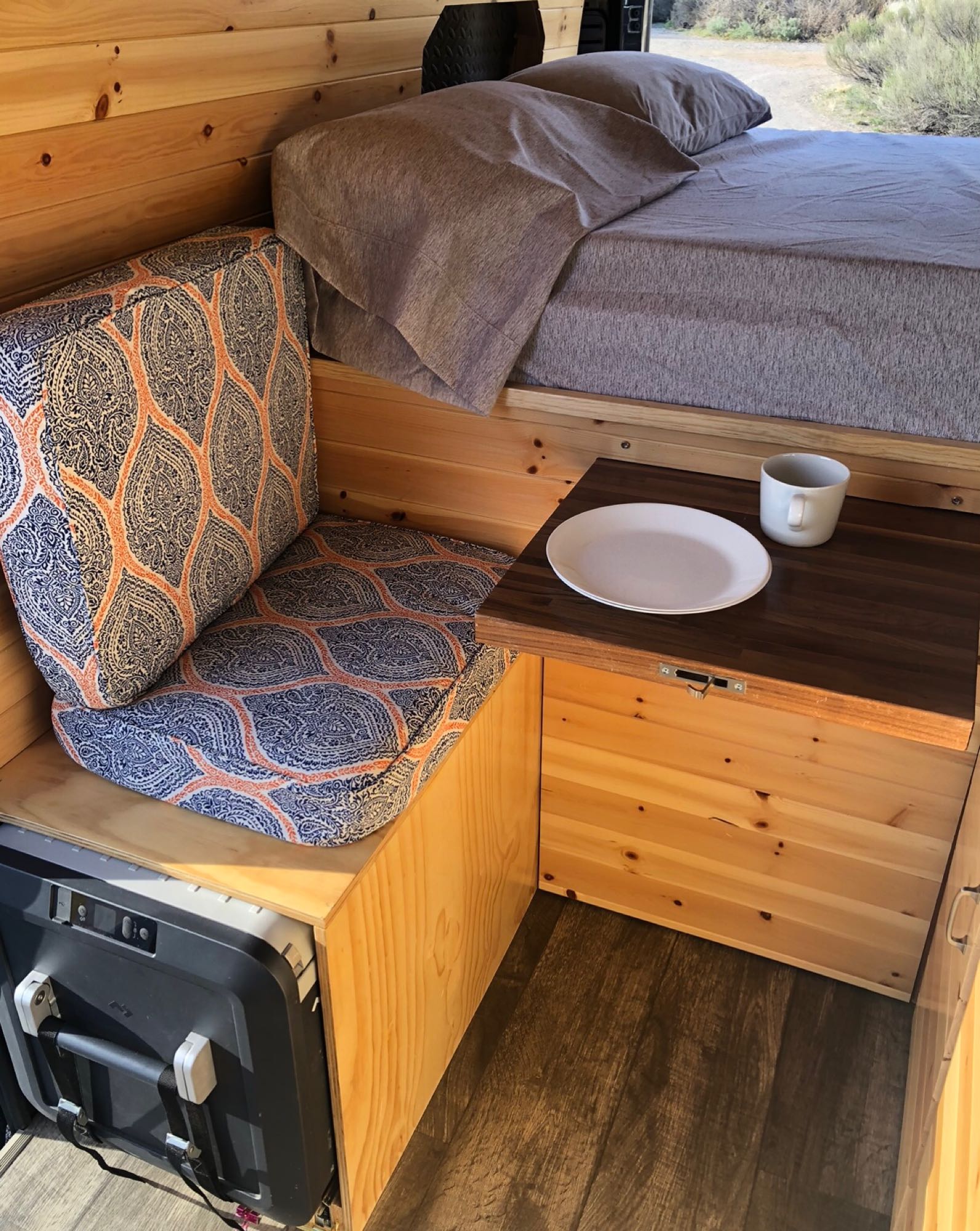 Cozy RV rental interior: wood walls, comfy bed, patterned seating, and a pull-out table with plate and mug. Ideal for adventures.. Dodge Ram 2500 2019