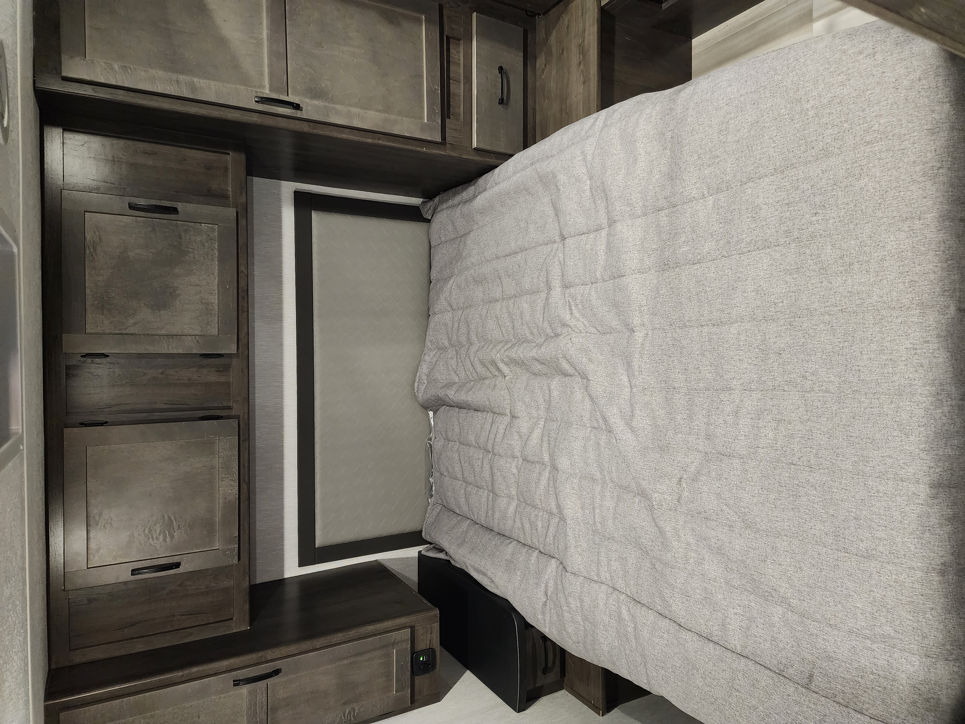 Cozy rv rental bedroom showing a bed with grey comforter and wall-mounted wooden storage cabinets.. Coachmen Leprechaun 2023