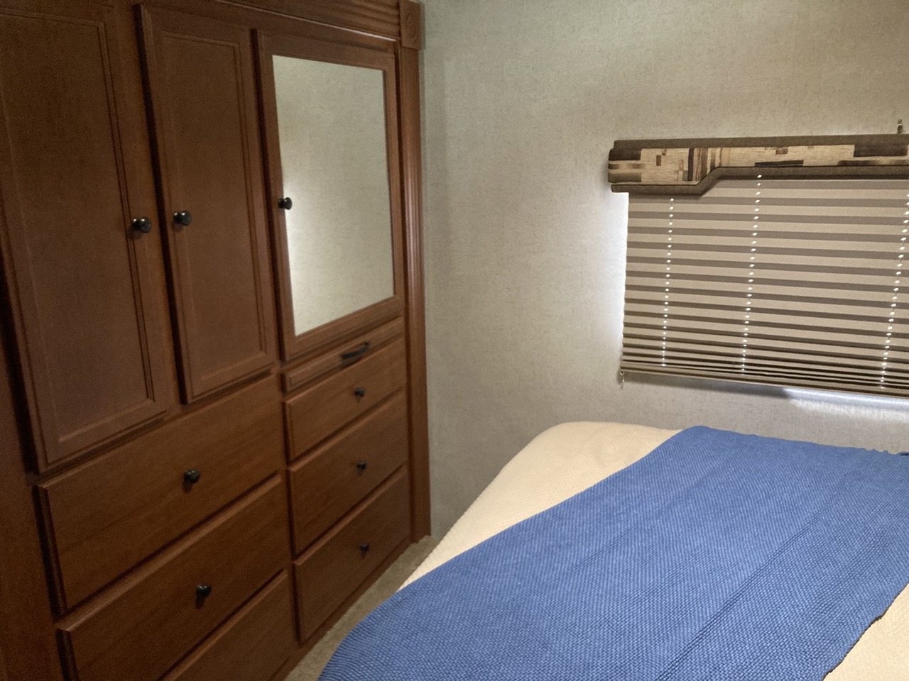 Compact RV rental bedroom showing a wooden wardrobe, mirror, multiple drawers, and bed with blue blanket under a pleated window blind.. Jamboree Jamboree Motorhome 2011