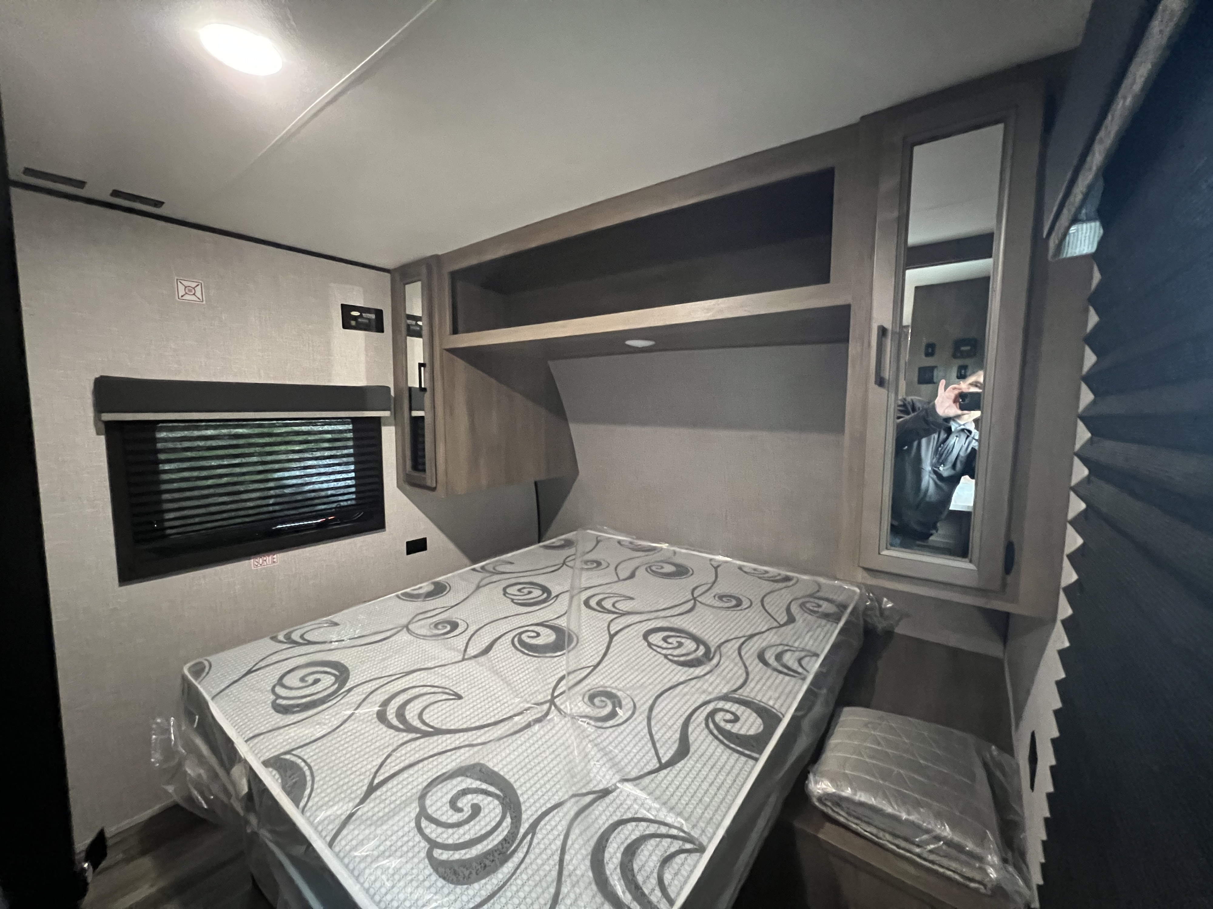 A new, plastic-wrapped mattress in an RV rental bedroom with overhead storage, a window with blinds, and a mirrored cabinet.. Starcraft Autumn Ridge 2023