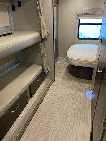RV rental interior: A functional camper space with twin bunk beds, a main bed, window, and storage, featuring light wood-patterned flooring.. Entegra Coach Emblem 2019