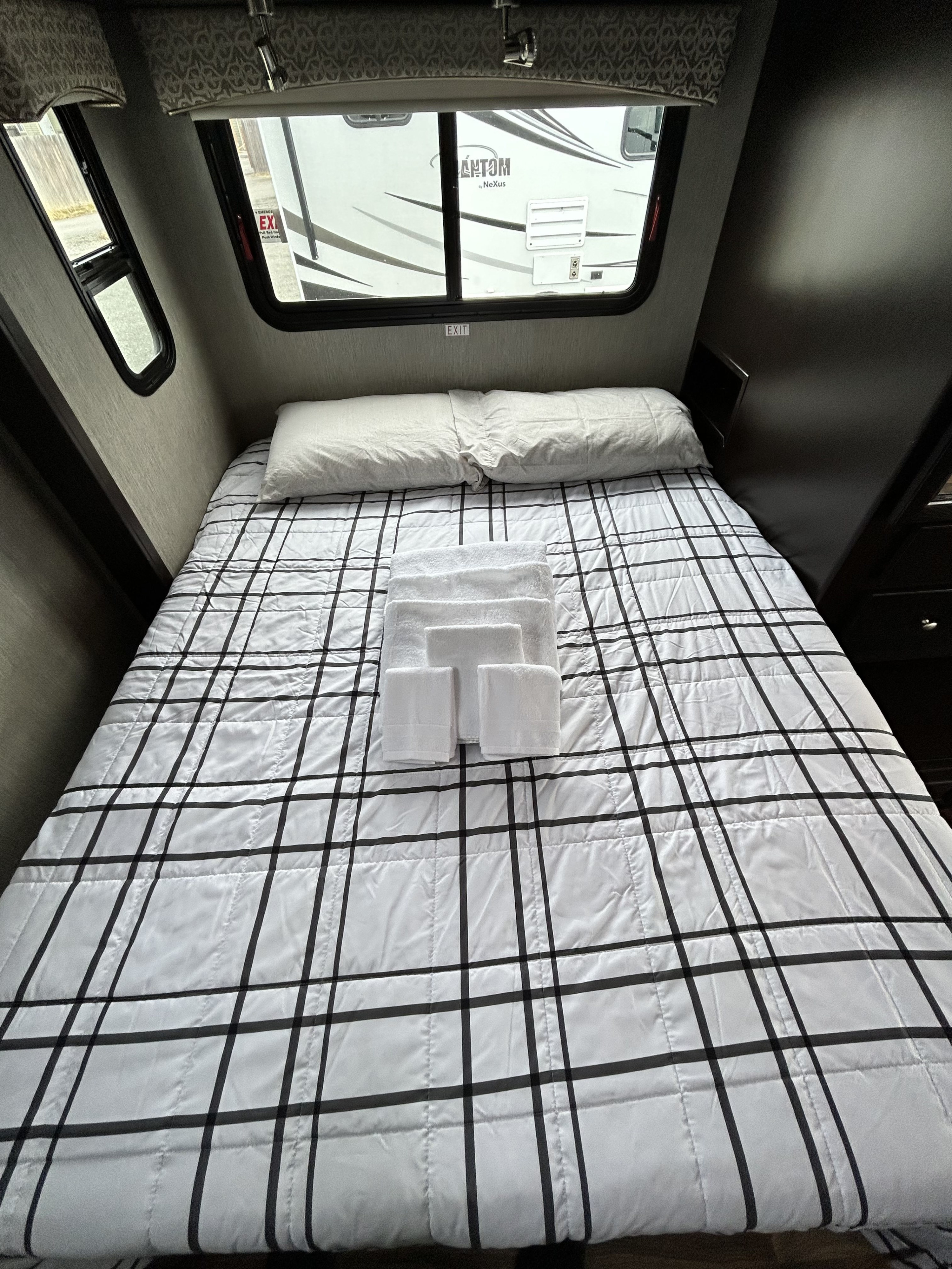 Cozy RV rental bedroom featuring a black grid bedspread, pillows, and fresh white towels for a comfortable journey.. Nexus Phantom 2020