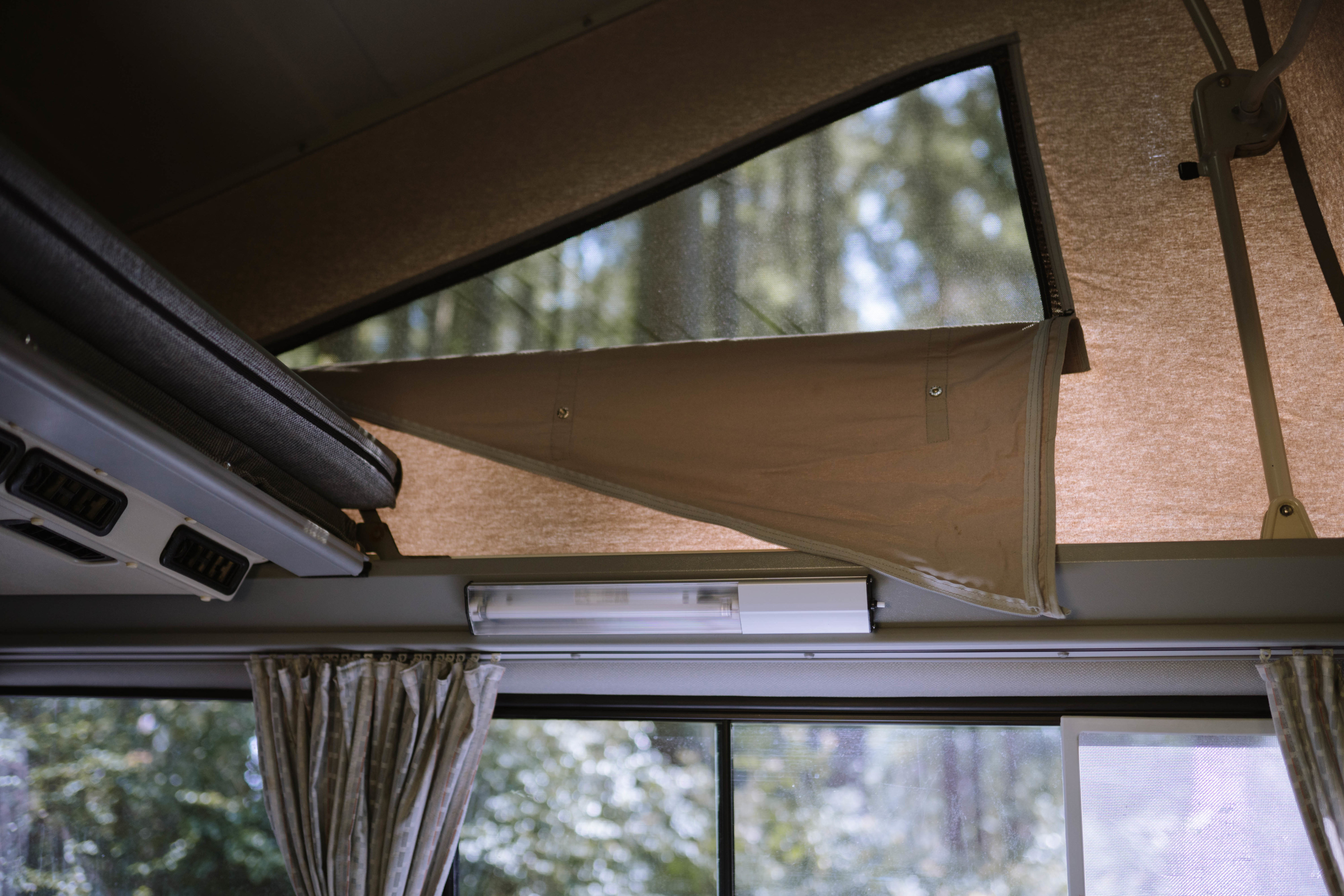 Inside an RV rental camper: a raised pop-top tent with a mesh window views of trees, and a lower window with curtains.. Volkswagen Westfalia 1990