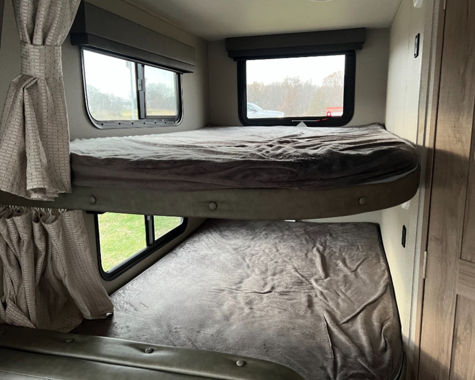 Inside an RV rental, showing two stacked bunk beds with soft blankets and windows overlooking nature. Cozy travel sleeping space.. Grand Design Imagine XLS 2021