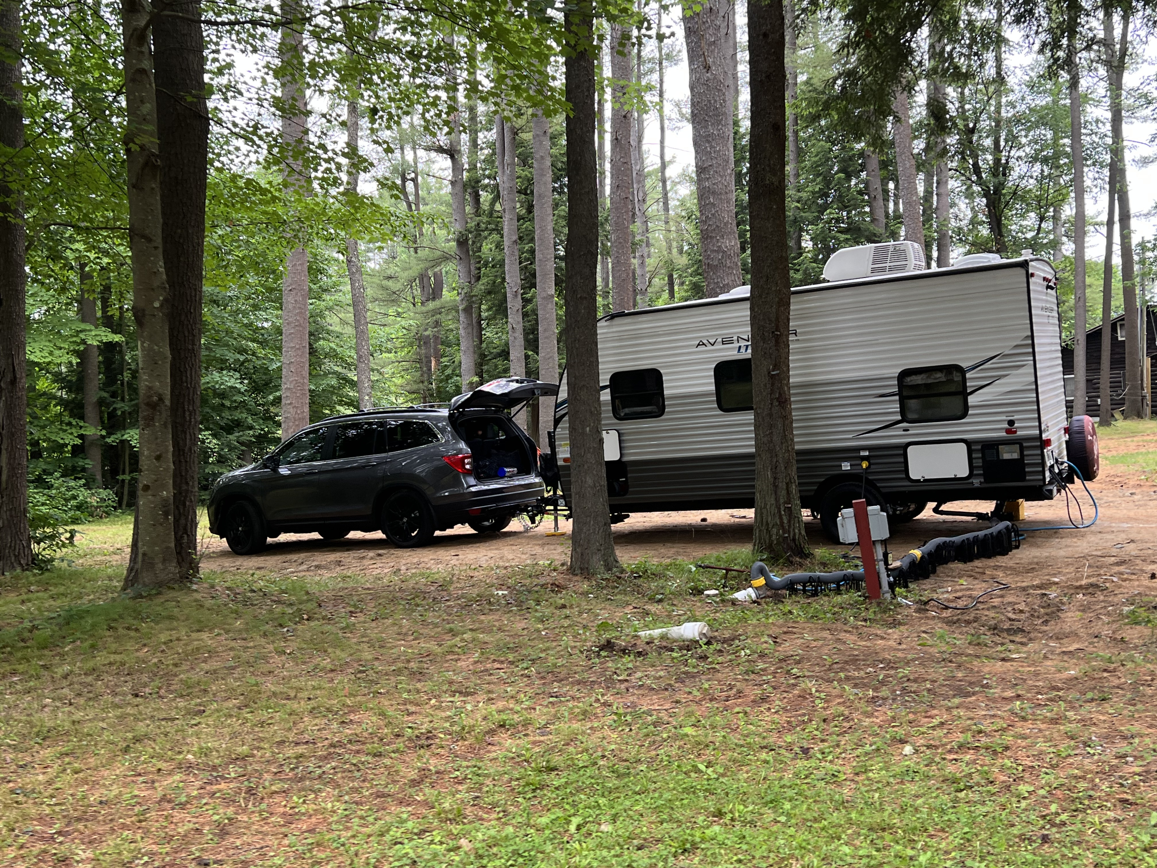 A grey SUV and an RV rental travel trailer are parked at a wooded campsite with utility hookups visible on the ground.. Avenger LT 2021