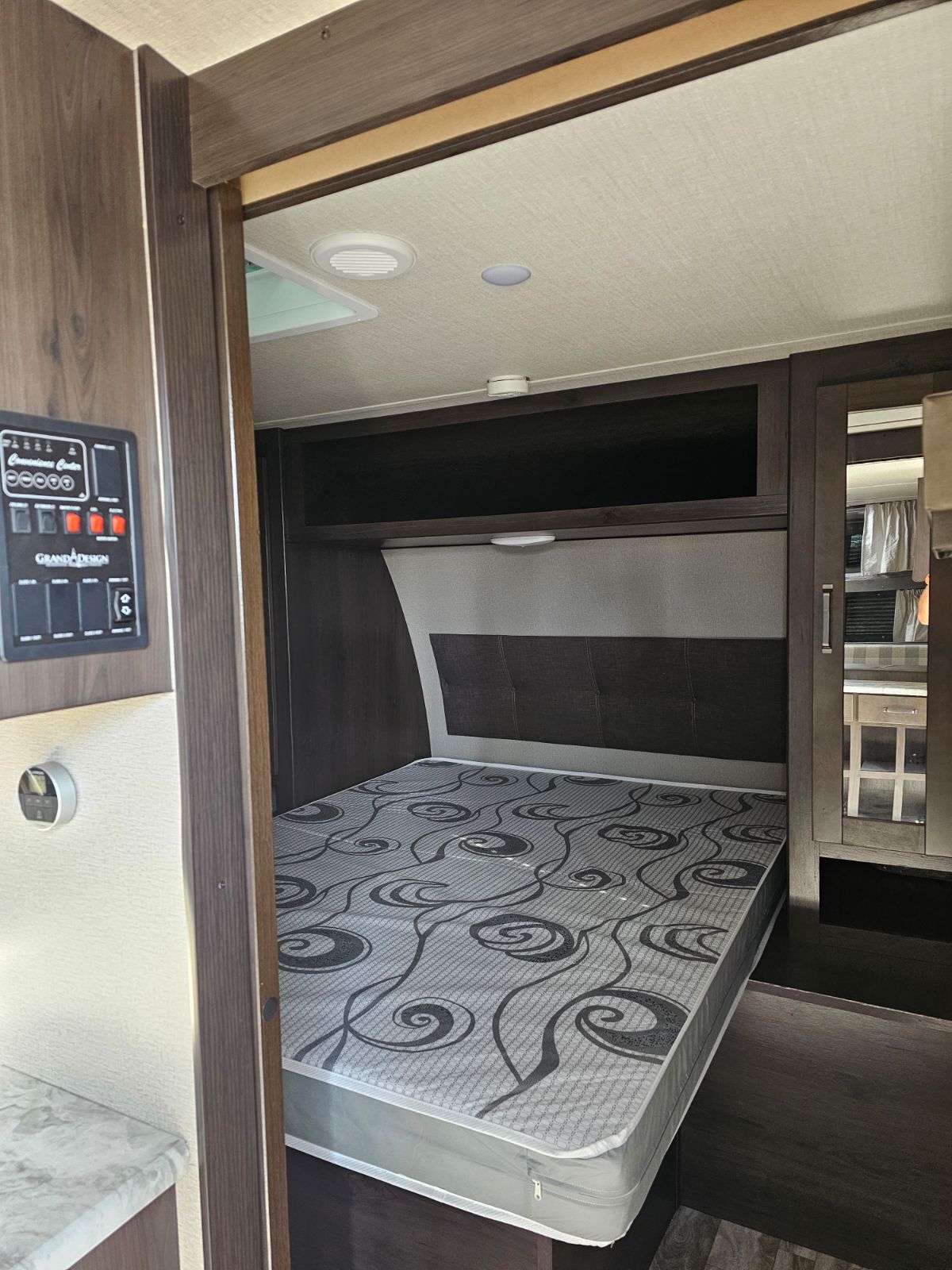 Inside an RV rental: a cozy bedroom features a gray patterned mattress, overhead storage, and a control panel nearby.. Grand Design Transcend Xplor 2022