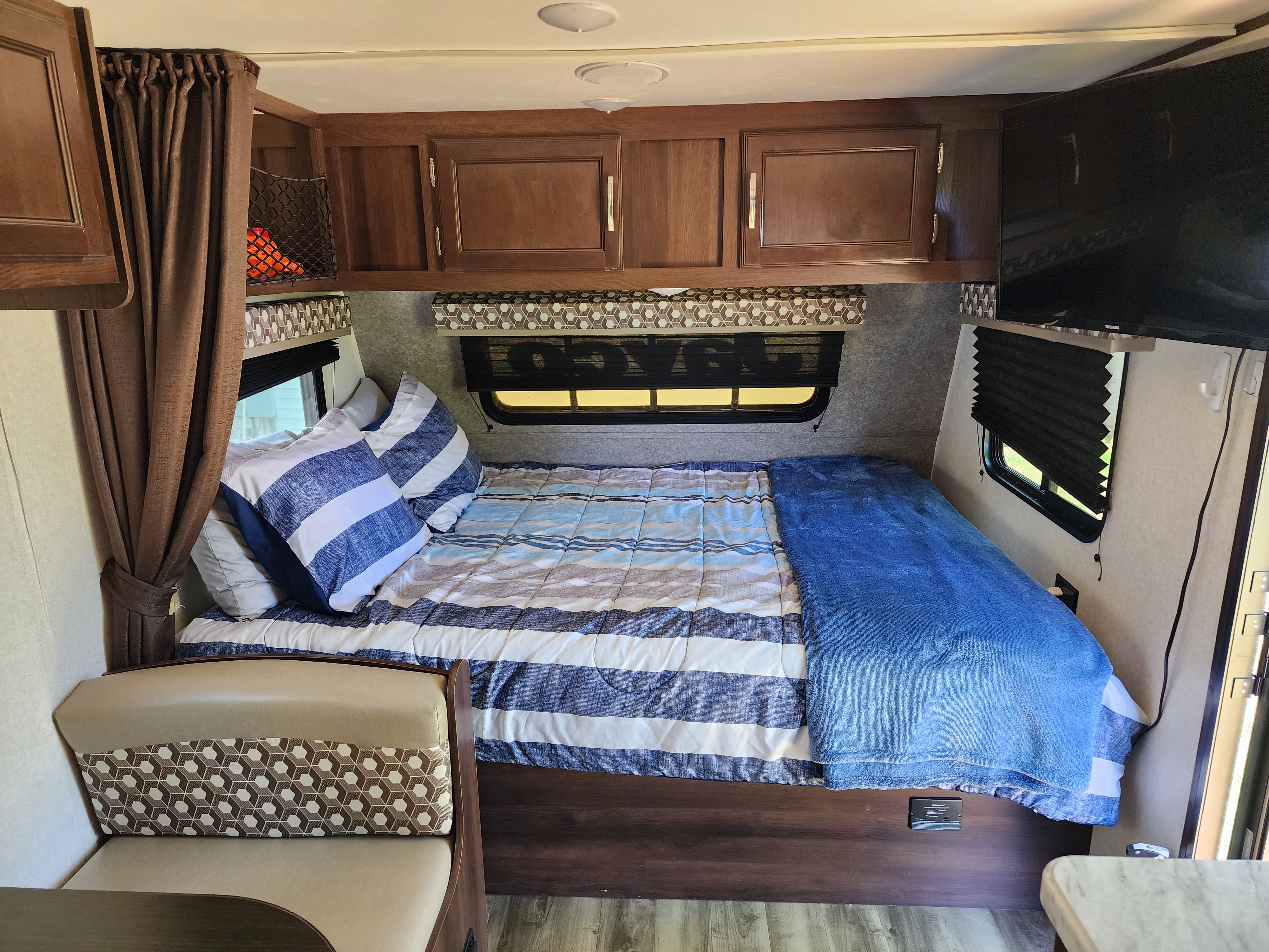 Cozy RV rental bedroom with blue and white striped bedding, overhead cabinets, and a small seating area.. Jayco Jay Flight SLX Baja Edition 2020