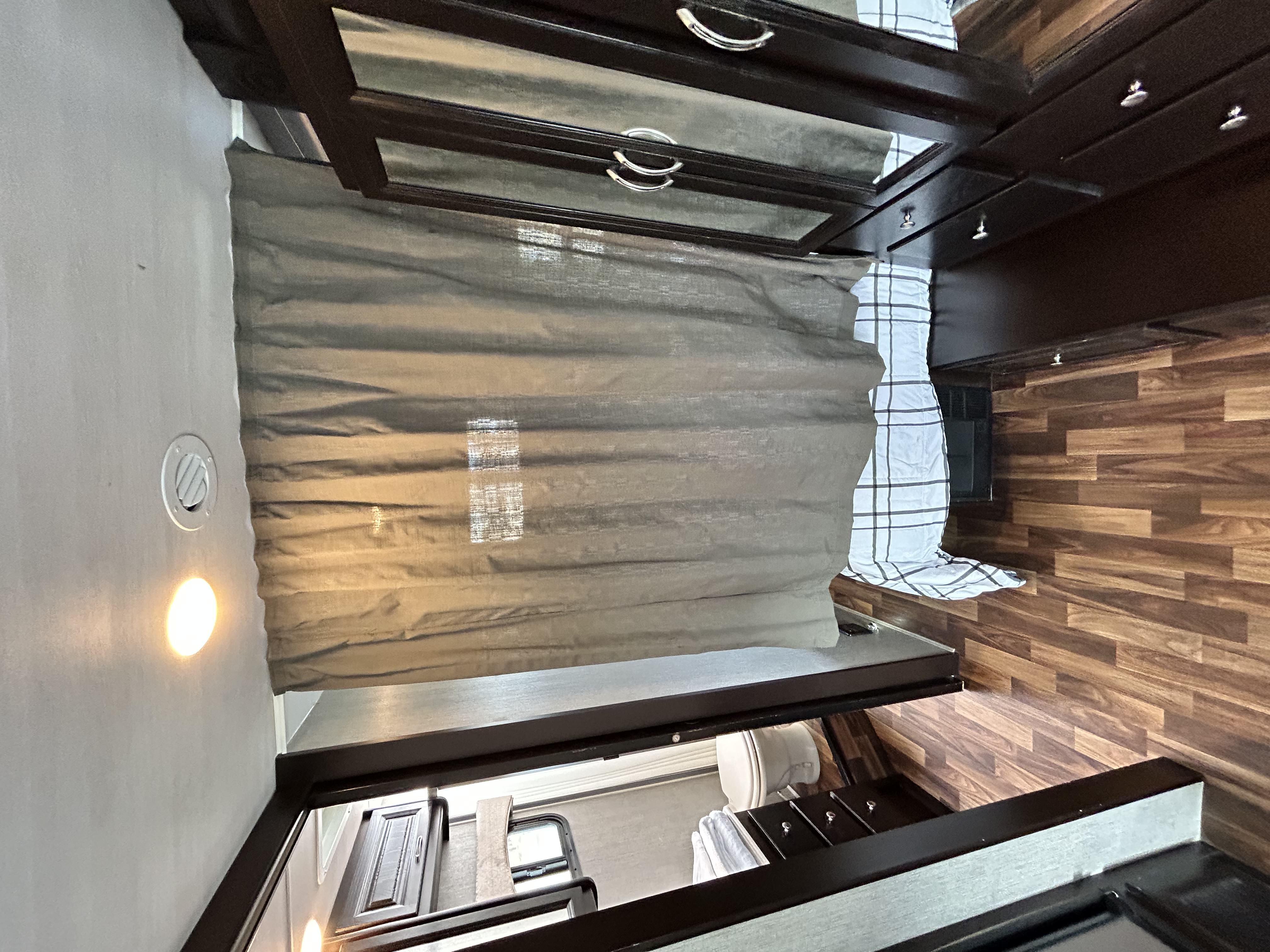 Bright RV rental interior with a cozy bed, dark cabinetry, and a pulled curtain, showcasing warm lighting and wood floors.. Nexus Phantom 2020