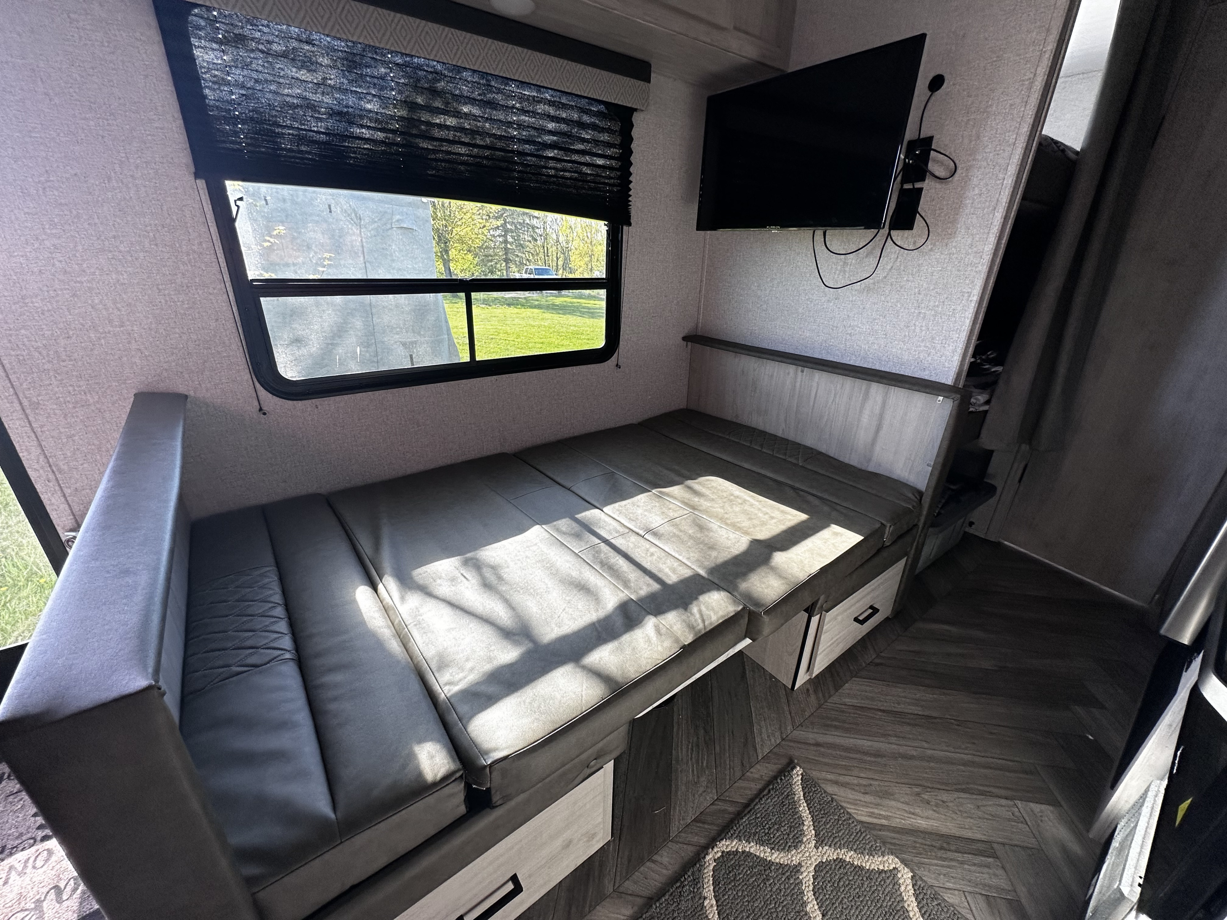 RV rental interior with a gray convertible bed, window view, storage, and wall-mounted TV.. East to West Della Terra 2022