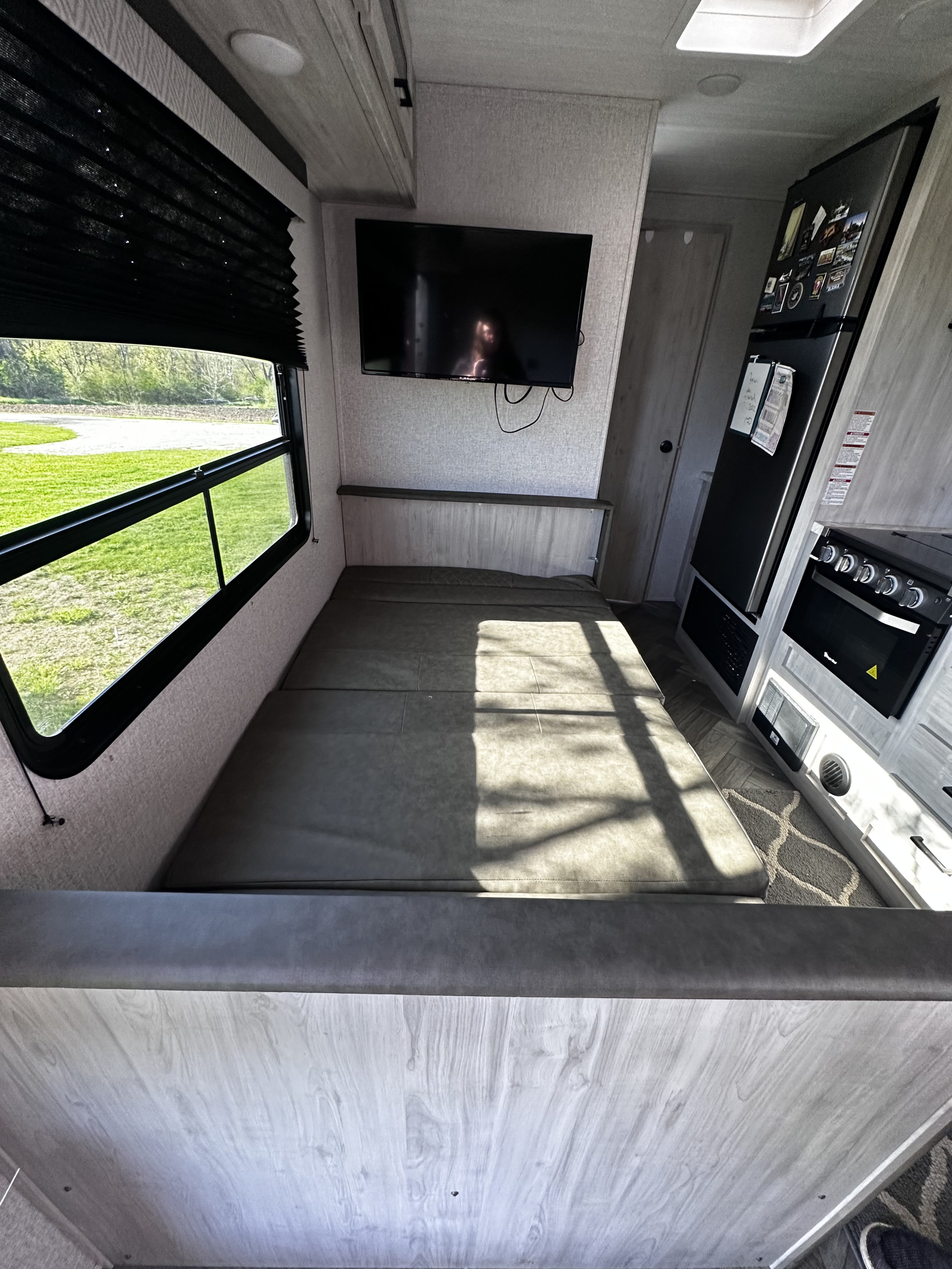 Comfortable RV rental interior showing a convertible bed, TV, kitchen area with fridge & stove, and a window overlooking nature.. East to West Della Terra 2022