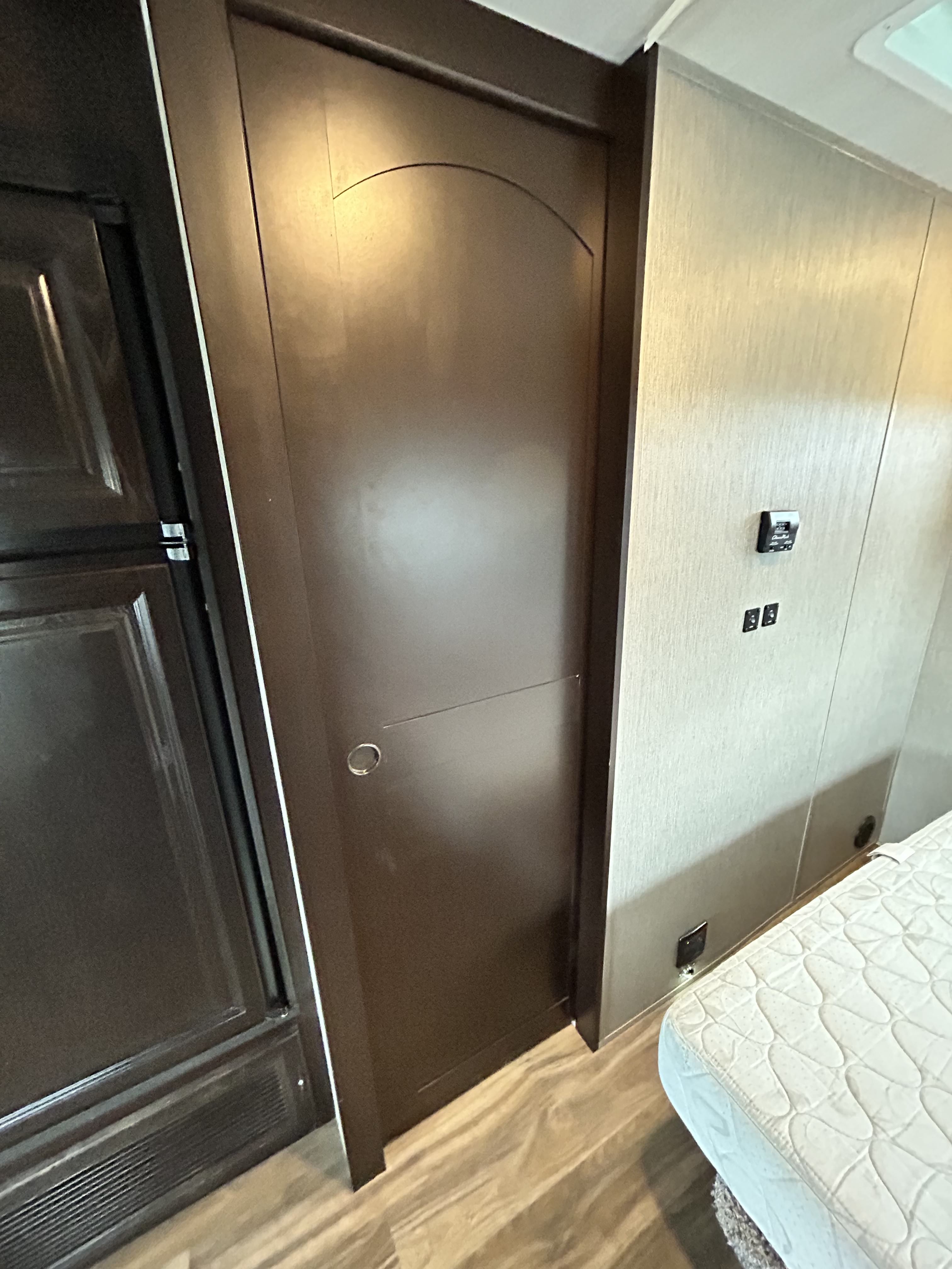 RV rental interior showing a dark wood door, part of a refrigerator, a white bed mattress, and wall-mounted controls.. Nexus Phantom 2020