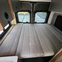 Comfortable grey upholstered bed in an RV rental camper van. The inviting sleeping area features large rear windows for light.. Winnebago Solis 2023