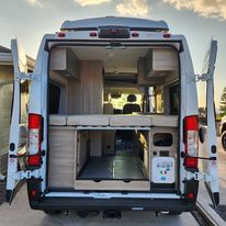 Open rear of a white camper van, showcasing a raised bed and spacious gear storage. Perfect for your next RV rental adventure.. Winnebago Solis 2023