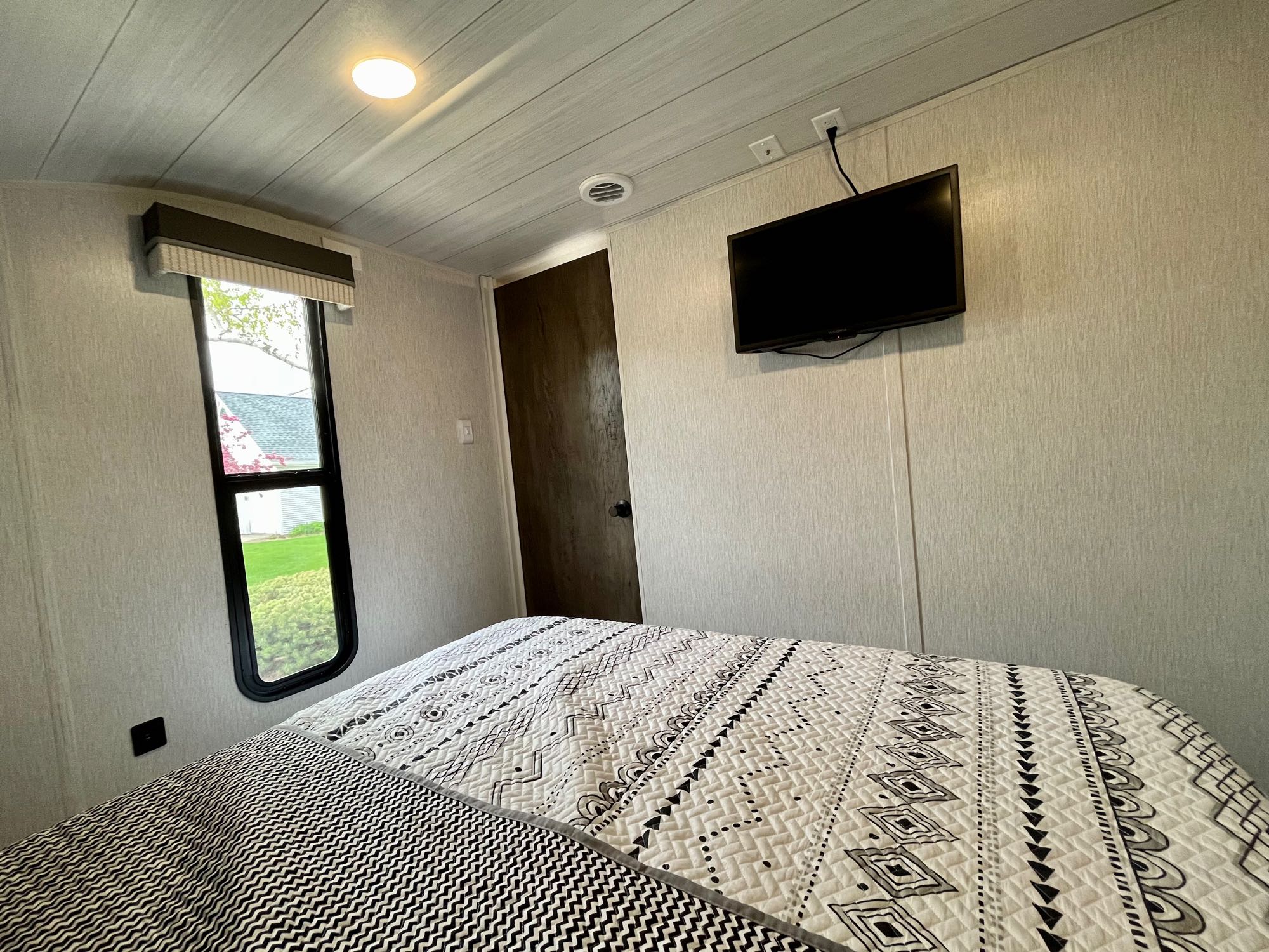 Inside a comfortable RV rental bedroom, showing a bed with a geometric quilt, wall-mounted TV, and window.. Forest River Salem Cruise Lite 2022