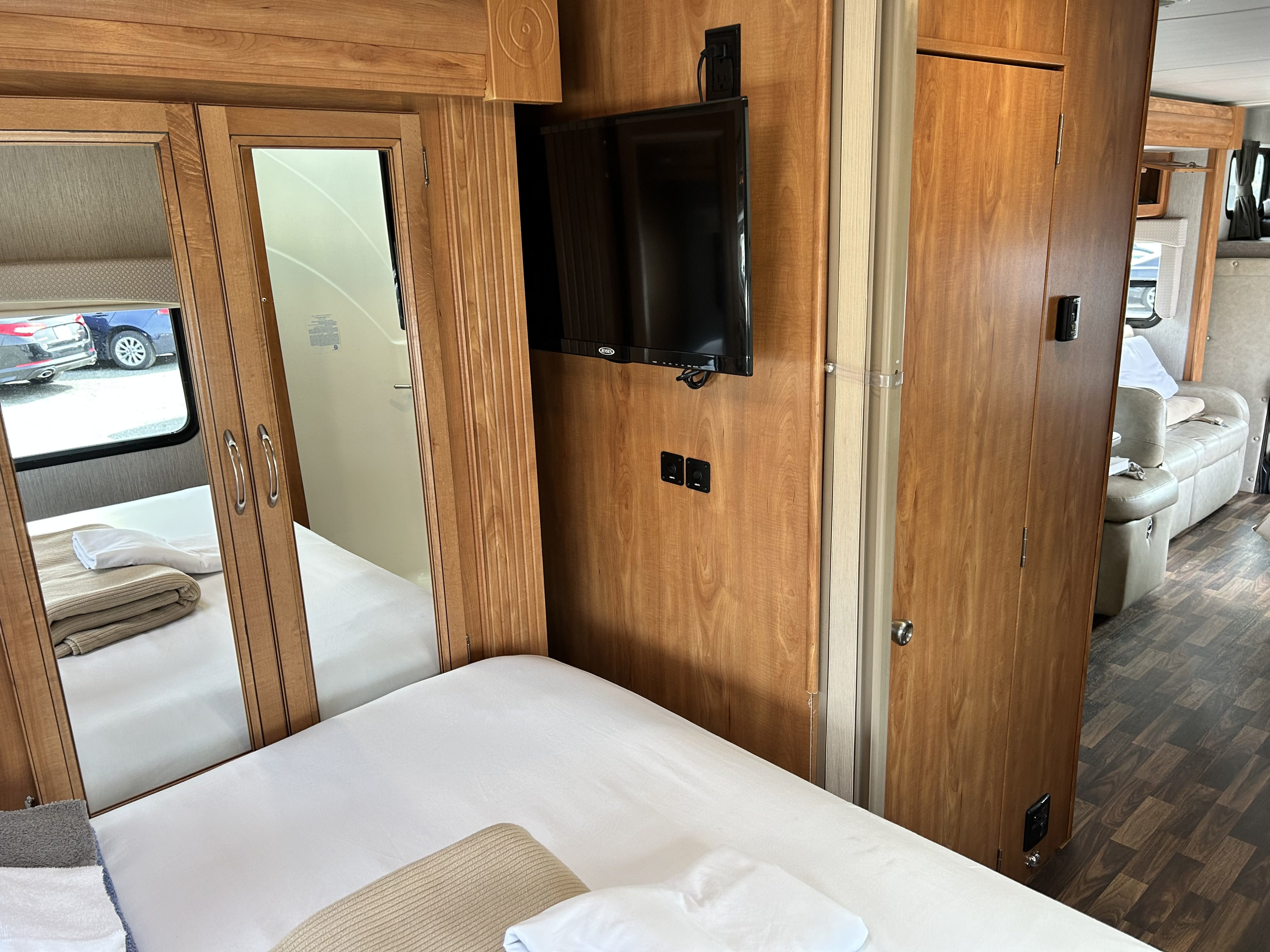 An RV rental bedroom offers a white bed, wall-mounted TV, mirrored closet doors, and a glimpse into the connecting lounge space.. Nexus Phantom 2019