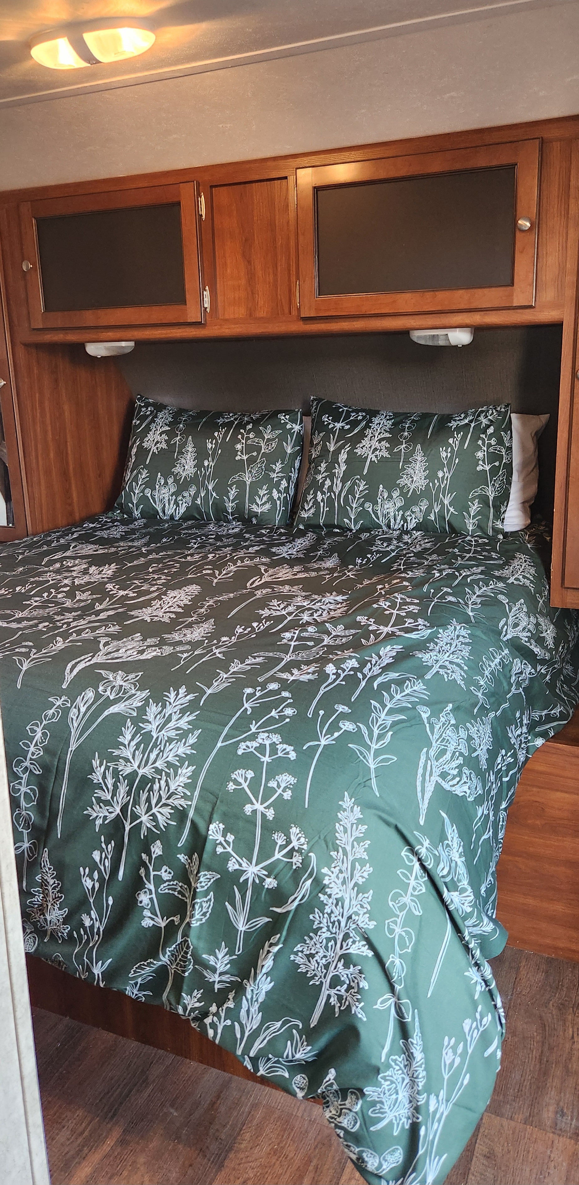 Serene RV rental bedroom. Features a bed with dark green botanical patterned bedding, under wooden overhead cabinets.. Keystone RV Bullet Ultra Lite 2015