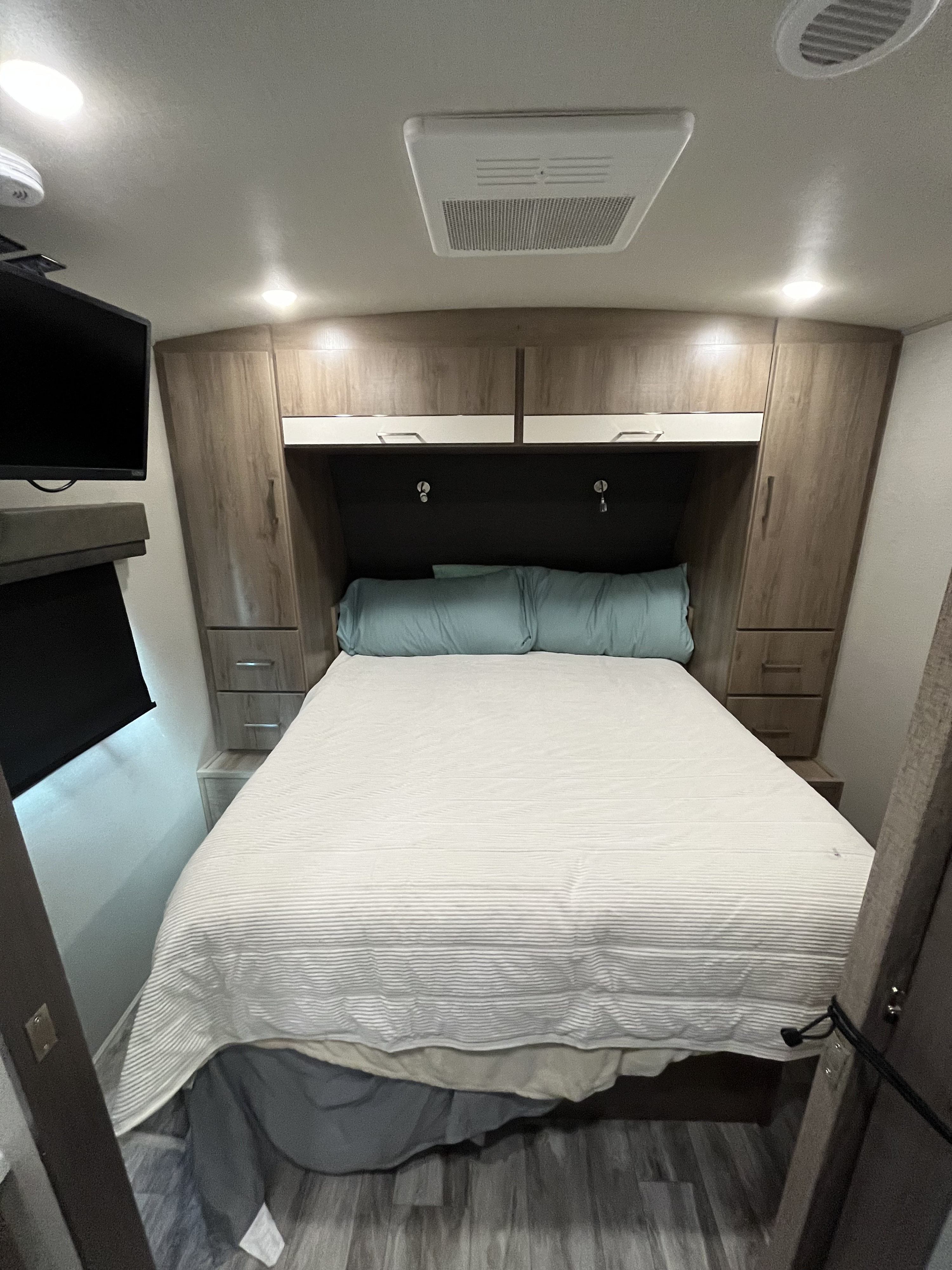 Compact RV rental bedroom with bed, overhead cabinets, TV, and AC unit. Wood flooring and light decor.. Grand Design Imagine 2022