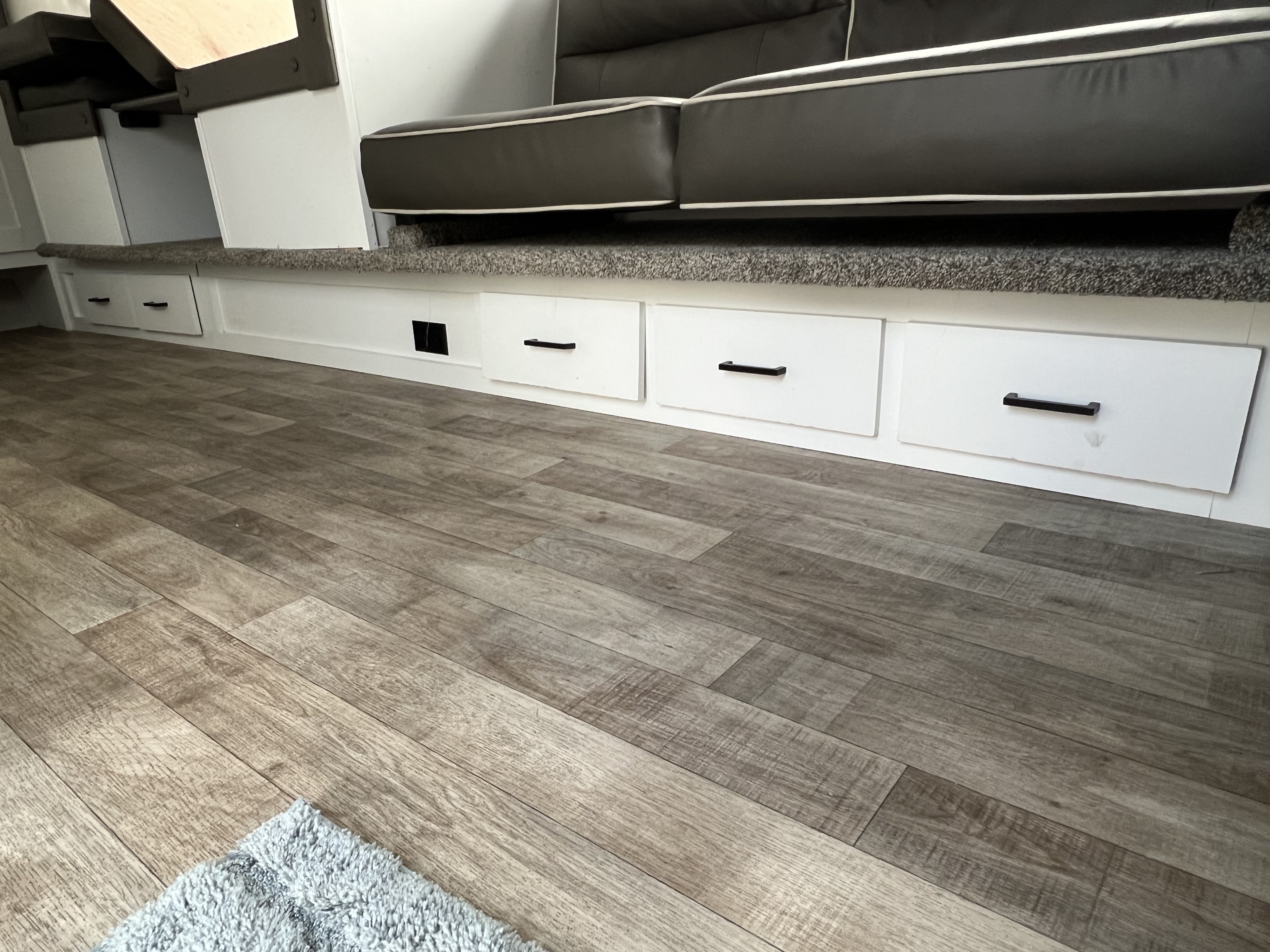 RV rental interior: wood-look floor, white storage drawers with black handles, and dark grey cushioned seating, possibly convertible.. Keystone Hideout 2021