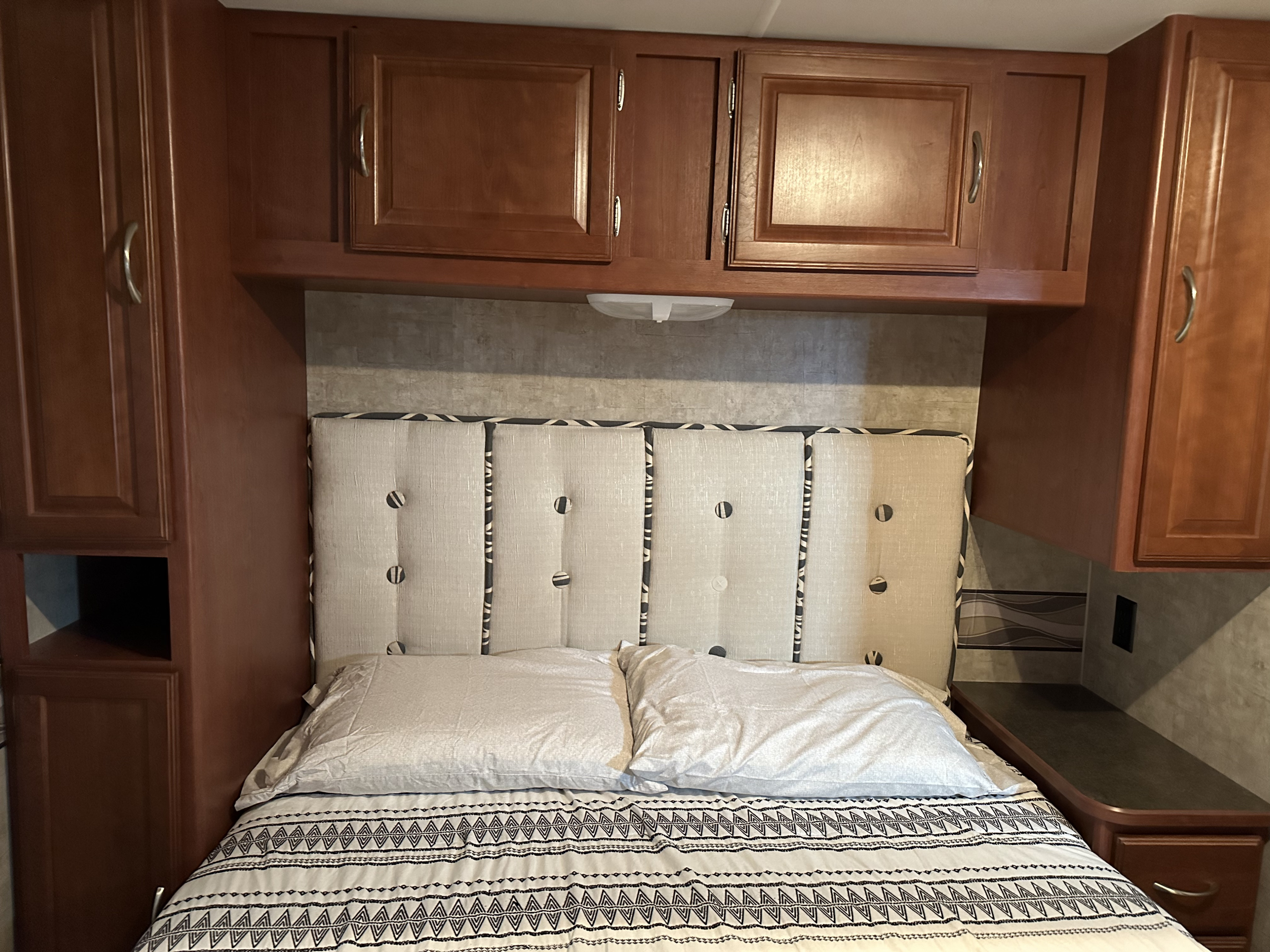 Cozy RV rental bedroom with a tufted headboard, wood cabinets, and bedside storage. Features a patterned duvet.. Winnebago Minnie Winnie 2015