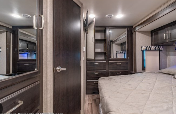 RV rental bedroom: A comfortable bed is visible on the right, surrounded by dark wood built-in storage with mirrors.. Entegra Coach Vision 2021