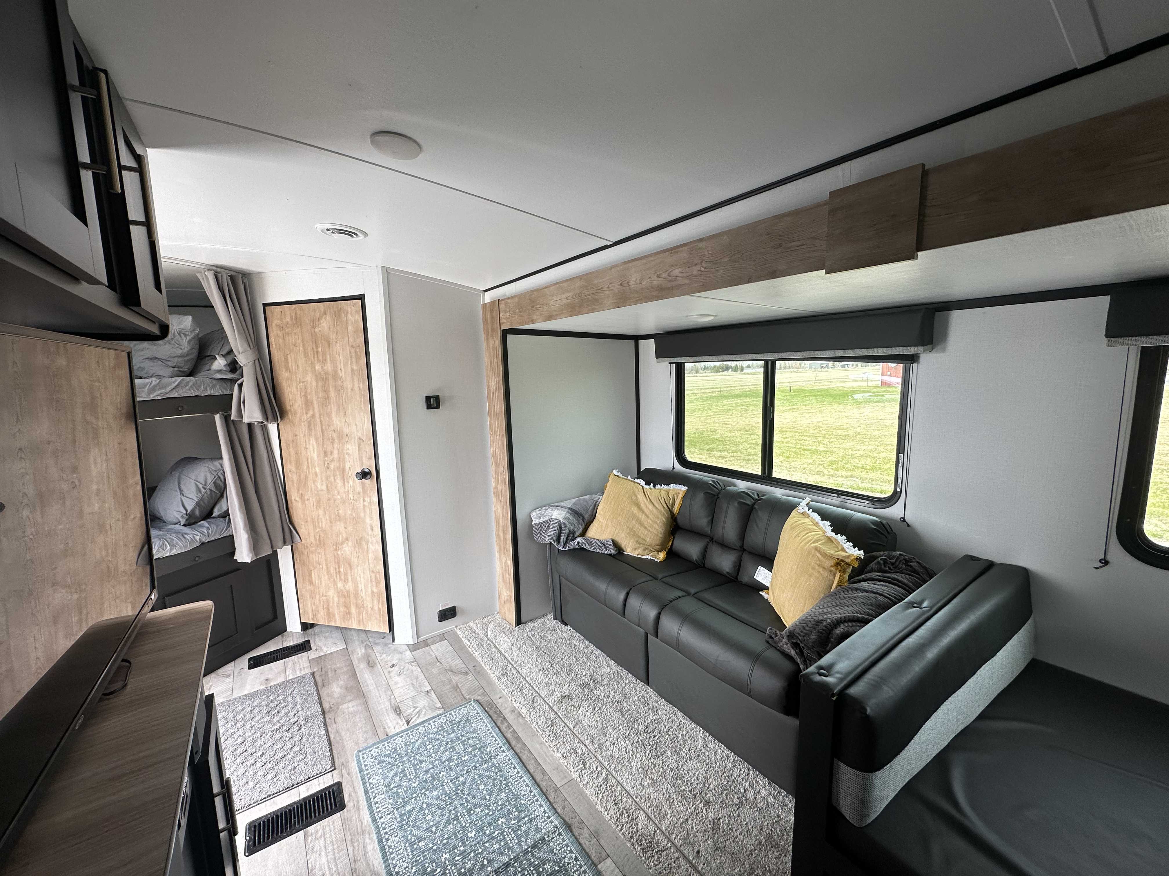 Inside this modern RV rental: a spacious living area with black sofa, bunk beds, and wide windows overlooking a green field.. Keystone RV Springdale 2022