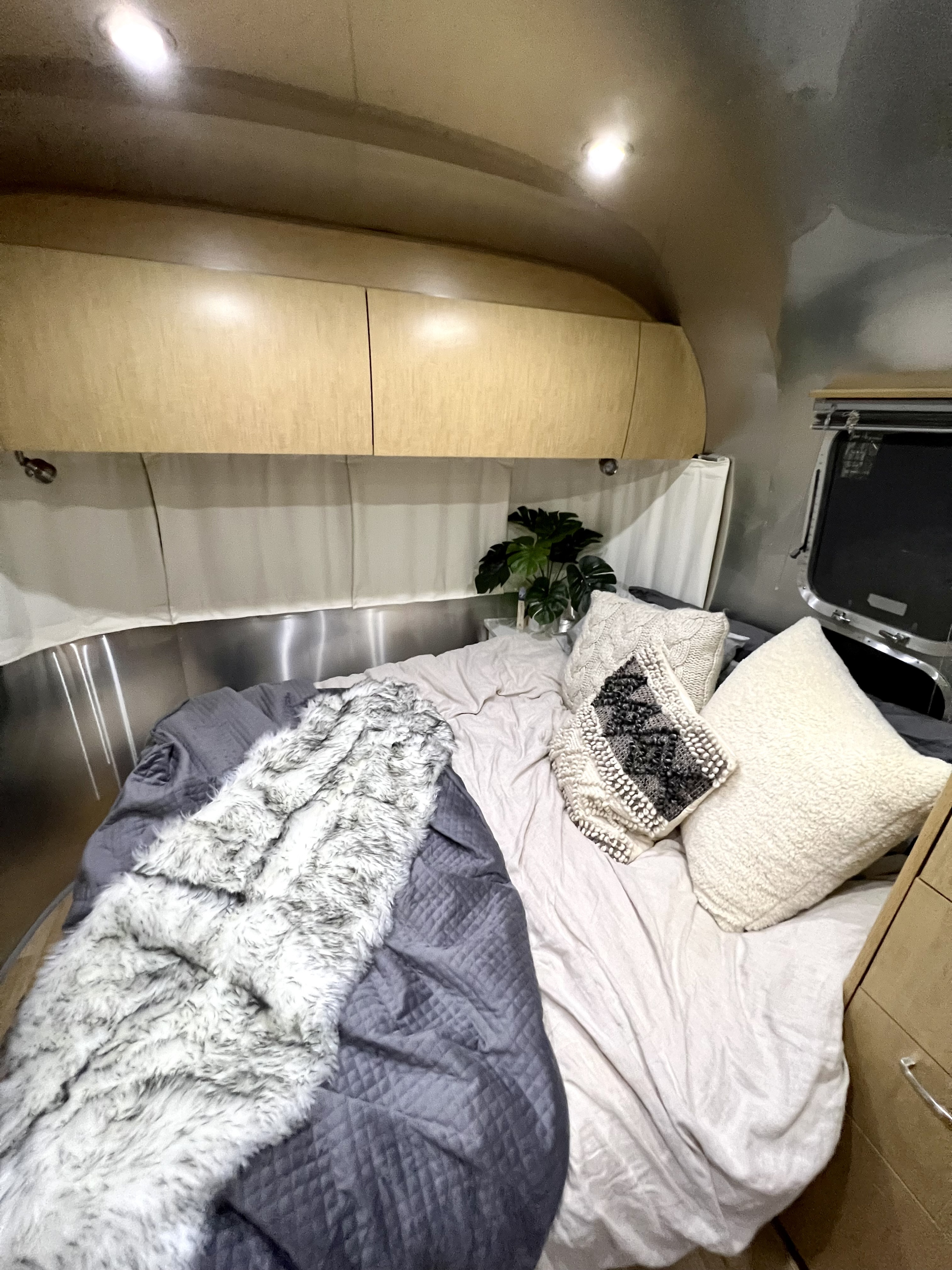 Inviting RV rental bedroom with soft lighting, light sheets, gray quilt, and a furry throw, plus wooden cabinets.. Airstream Flying Cloud 2014