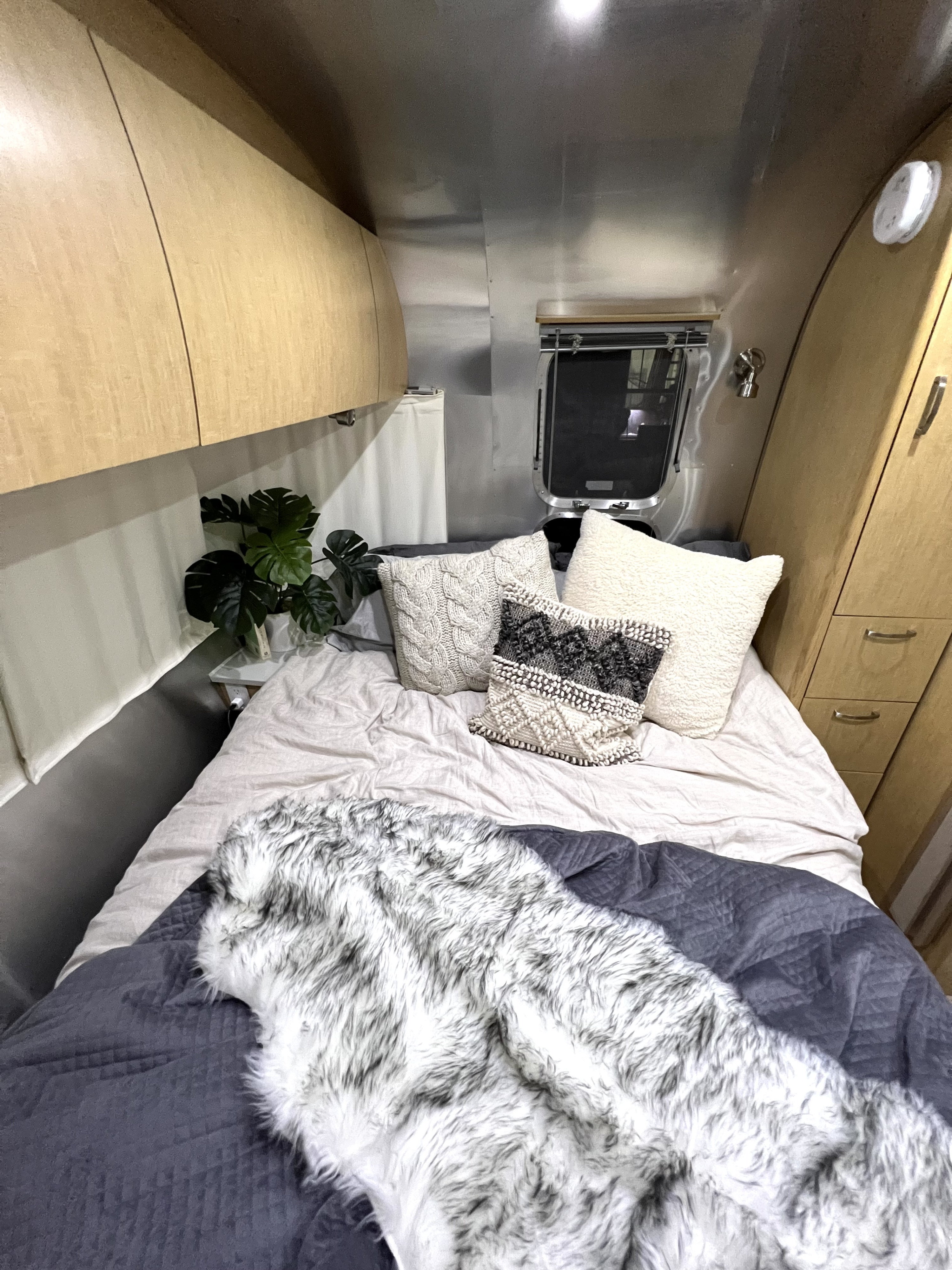 Cozy RV rental bedroom featuring a plush bed with textured pillows, a faux fur blanket, and overhead storage.. Airstream Flying Cloud 2014