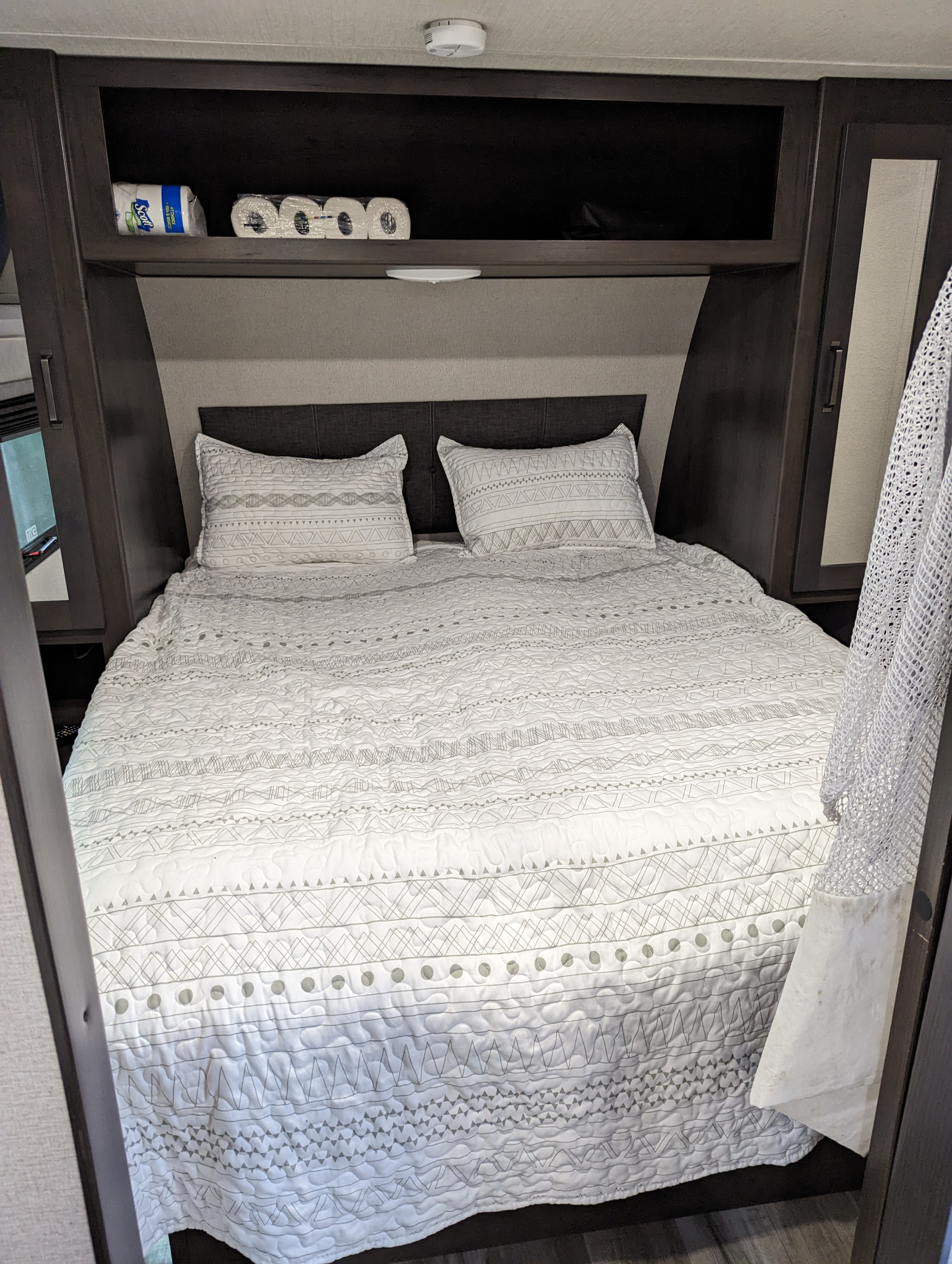 Experience comfort in this RV rental's bedroom, featuring a neatly made bed with patterned white bedding and ample overhead storage.. Grand Design Transcend Xplor 2022