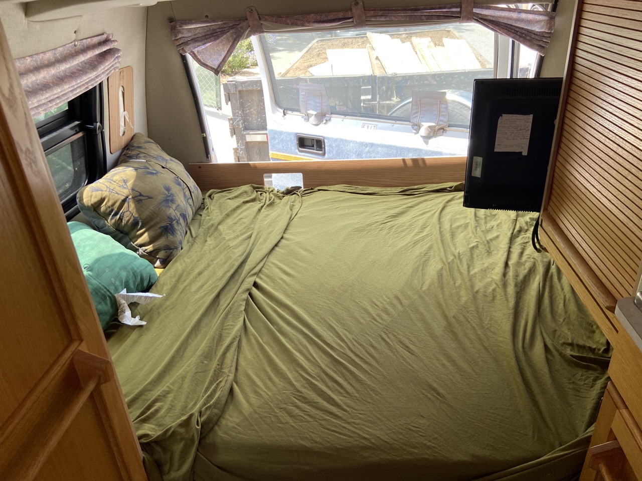 Interior of an RV rental: a neatly made bed with olive green sheets, pillows, and a mounted TV for comfy travel.. Roadtrek 190 Versatile 2003