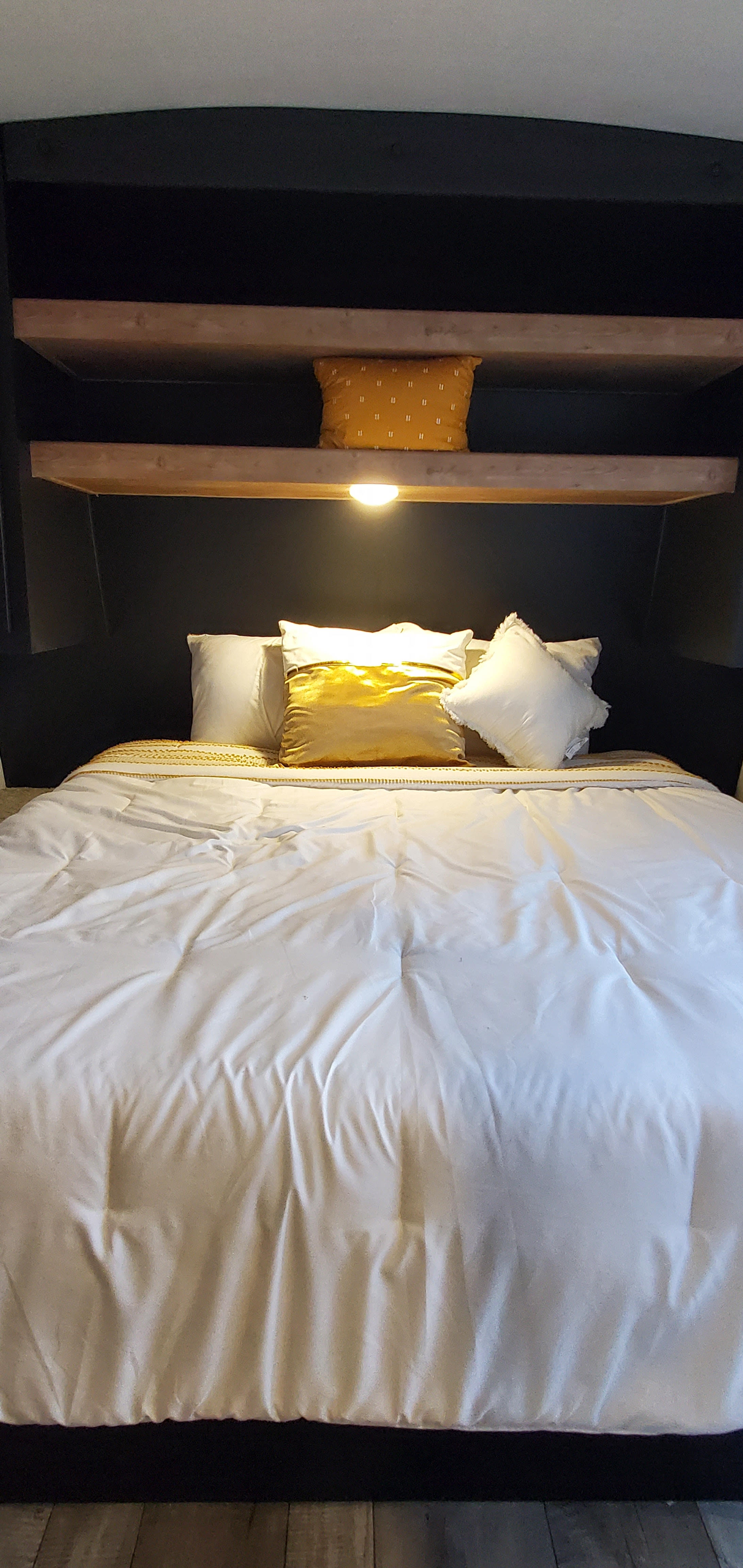 Inviting RV rental bed with white and gold pillows, clean white bedding, and built-in wooden shelves with lighting.. Keystone RV Springdale 2022