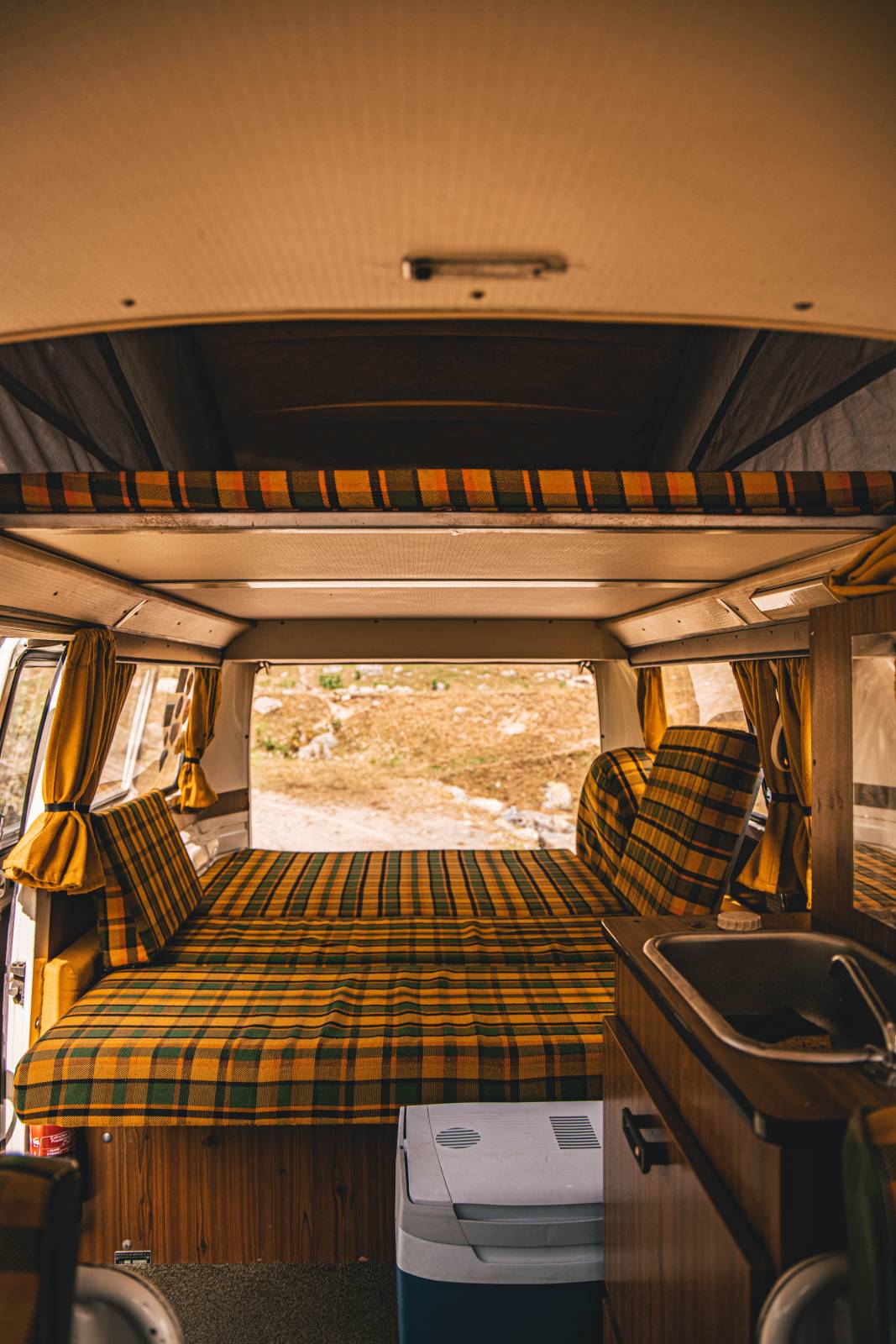 Explore this cozy RV rental interior featuring a comfy plaid bed, sink, and cooler – your perfect road trip home.. Volkswagen T2 Westfalia 1976