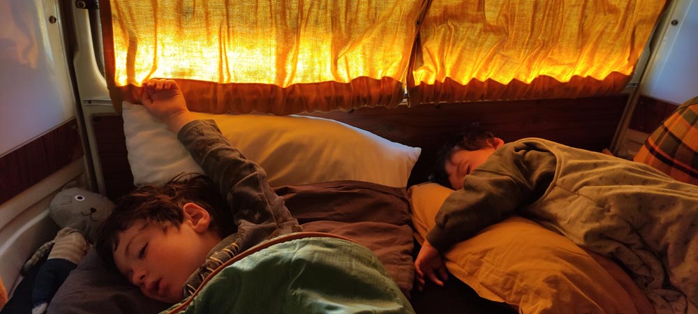 Cozy RV rental adventure: Two kids sound asleep in a warm camper, bright curtains filtering sunlight. Perfect family travel comfort.. Volkswagen T2 Westfalia 1976