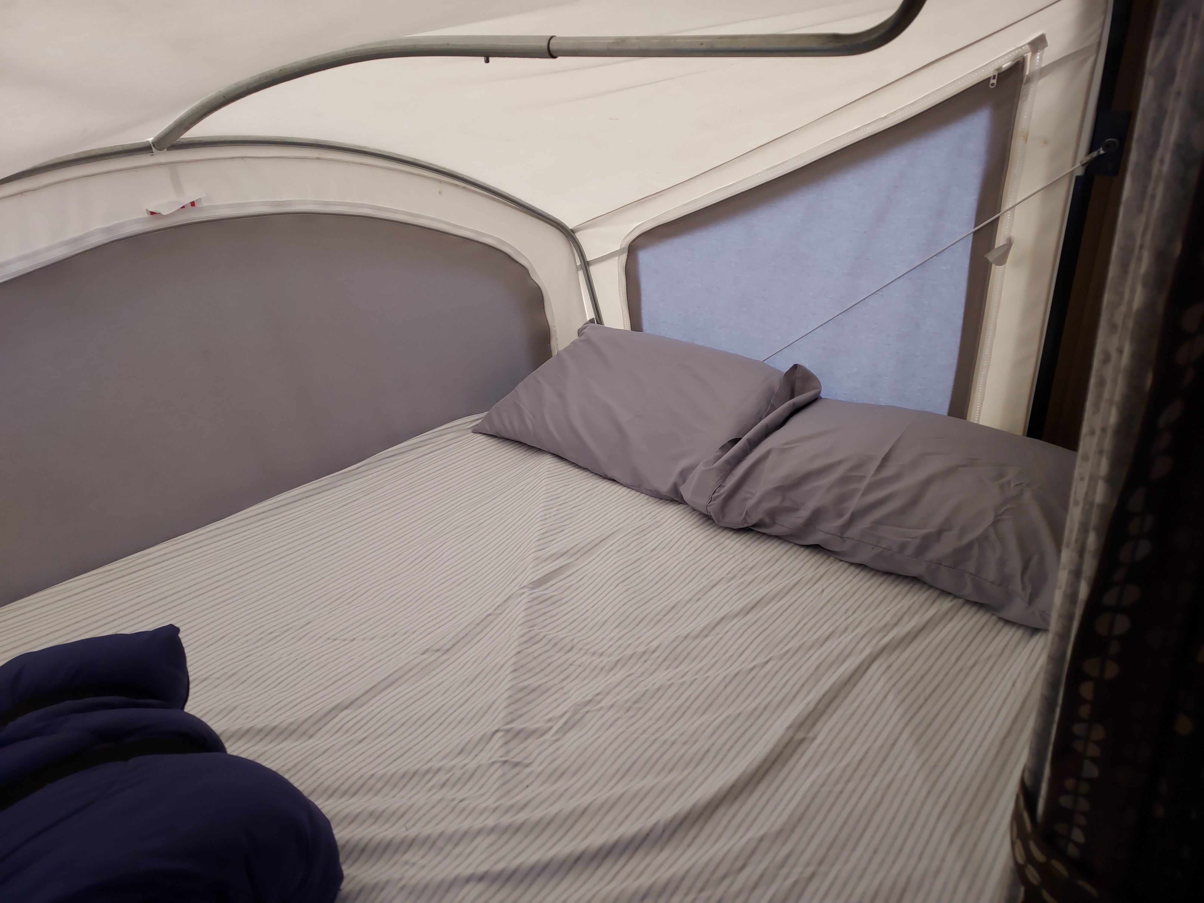 RV rental interior: a cozy bed with striped sheets, two grey pillows, and a window with a pull-down shade.. KZ Spree Escape Model 18RBT 2015