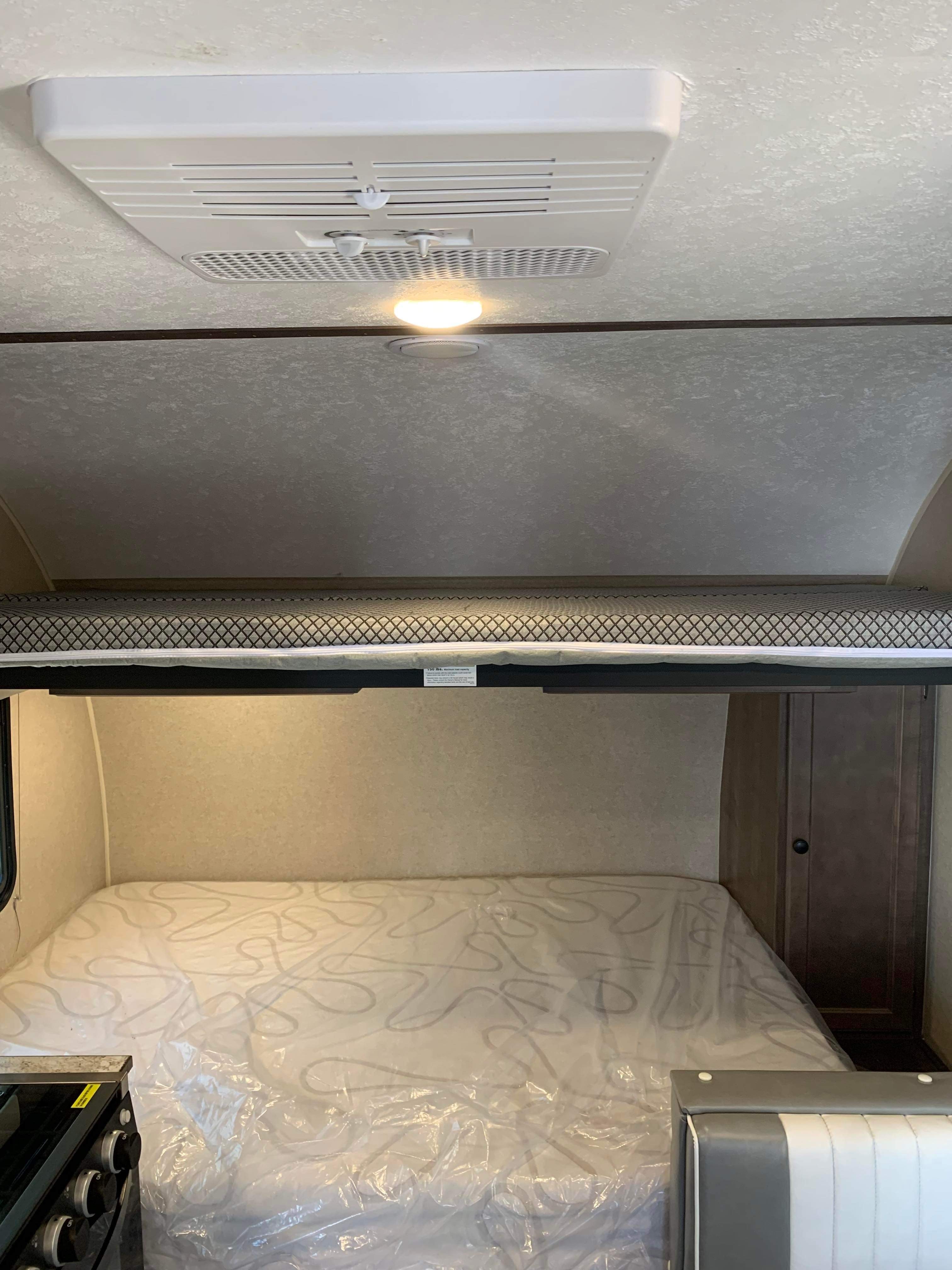 RV rental interior featuring an overhead AC unit, a bunk bed above, and a new, plastic-covered main bed area.. Sunset Park & Rv Inc. 16BH 2022